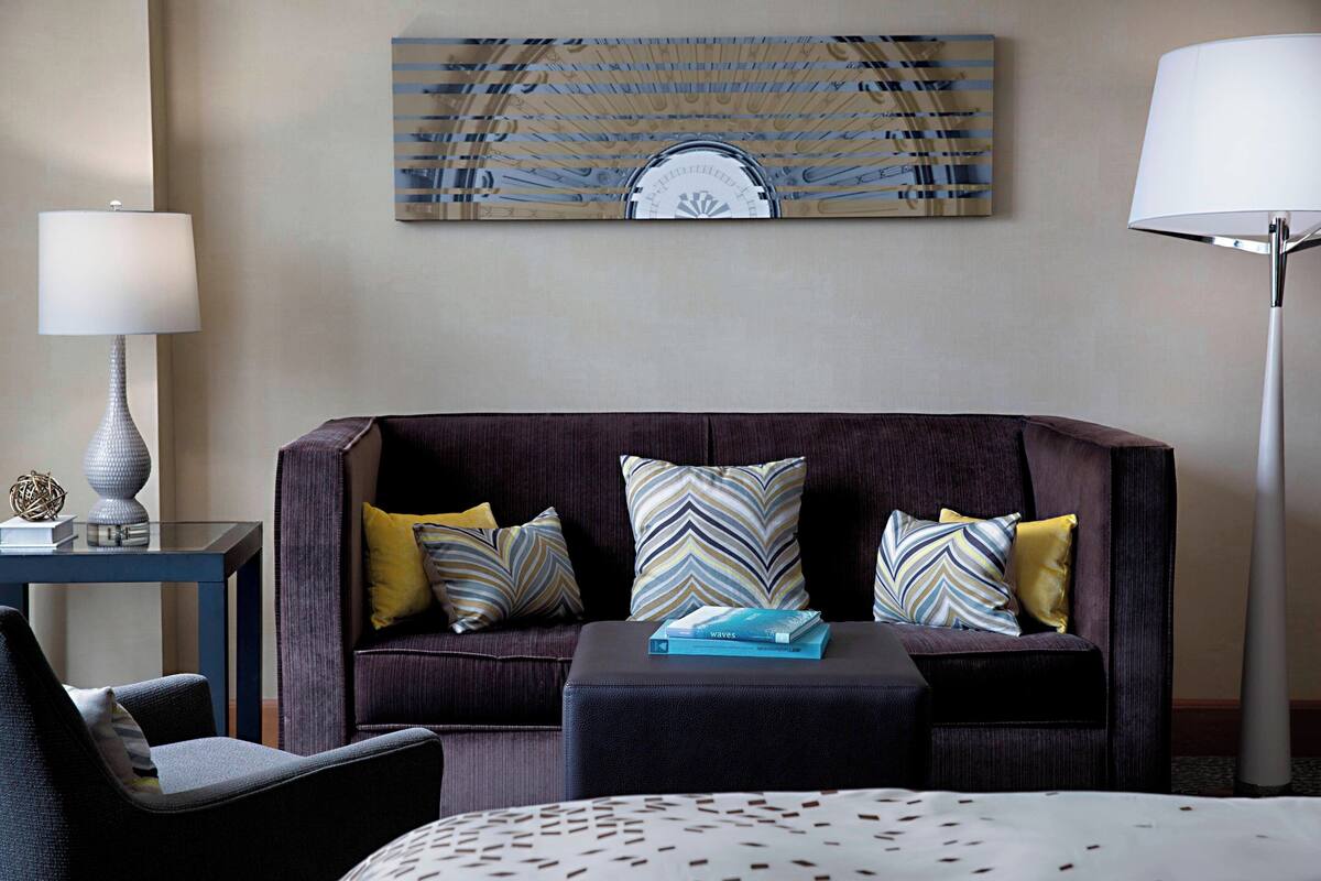 A hotel room with a dark brown sofa, two patterned pillows, a small table with a lamp, and a decorative wall piece above the sofa.