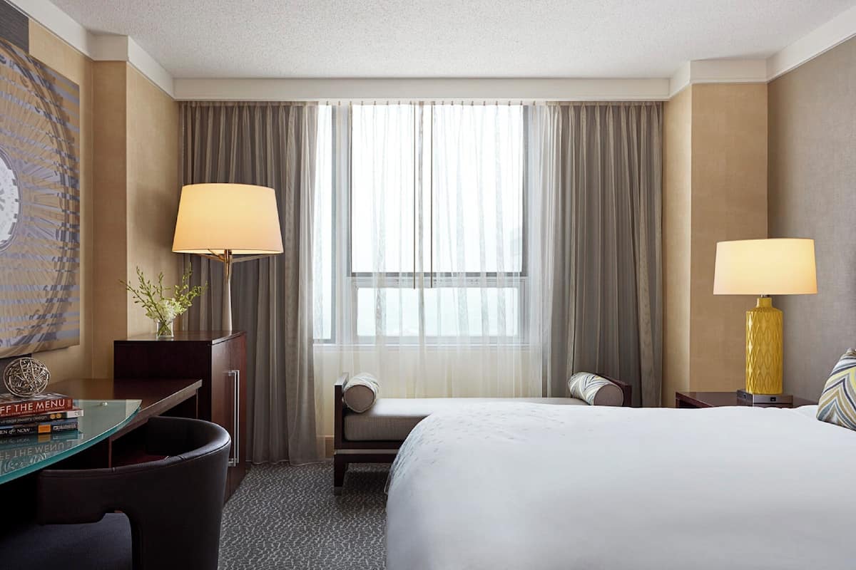 A hotel room with a large bed, two bedside lamps, a desk with a lamp, a chair, a small table with books, and a window with curtains.