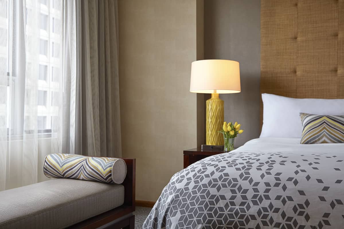 A hotel room with a bed, a nightstand with a lamp, a chair, and a window with curtains.