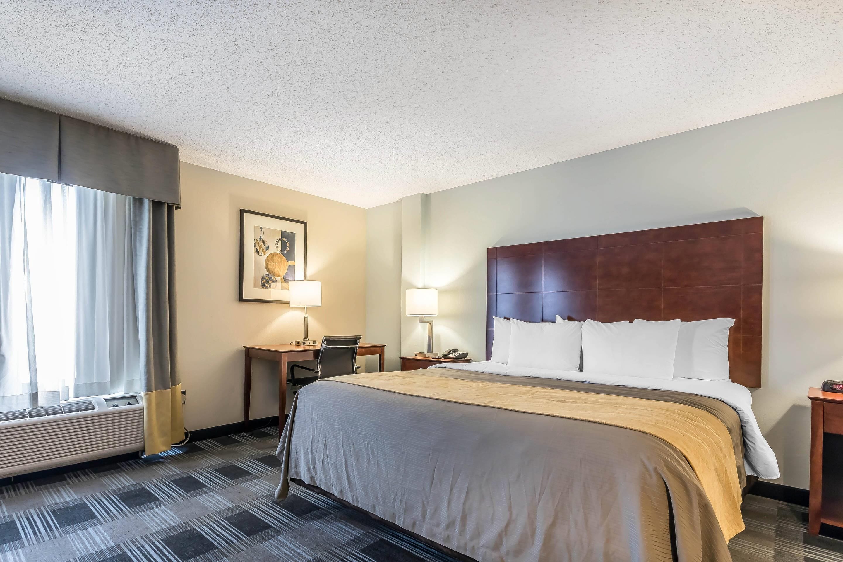 Una habitación de hotel con una cama grande, un escritorio, dos lámparas, un cuadro en la pared y una ventana con cortinas.