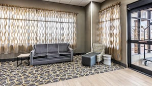 A hotel room with a patterned carpet, a gray sofa, a small table, a patterned armchair, and a large window with curtains.