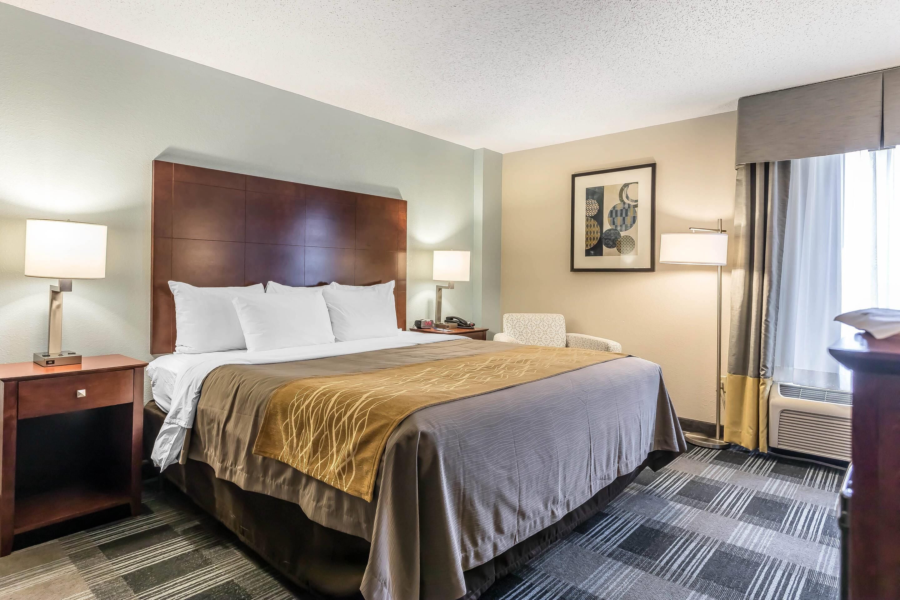 A hotel room with a large bed, two bedside tables with lamps, a chair, a framed artwork on the wall, and a window with curtains.