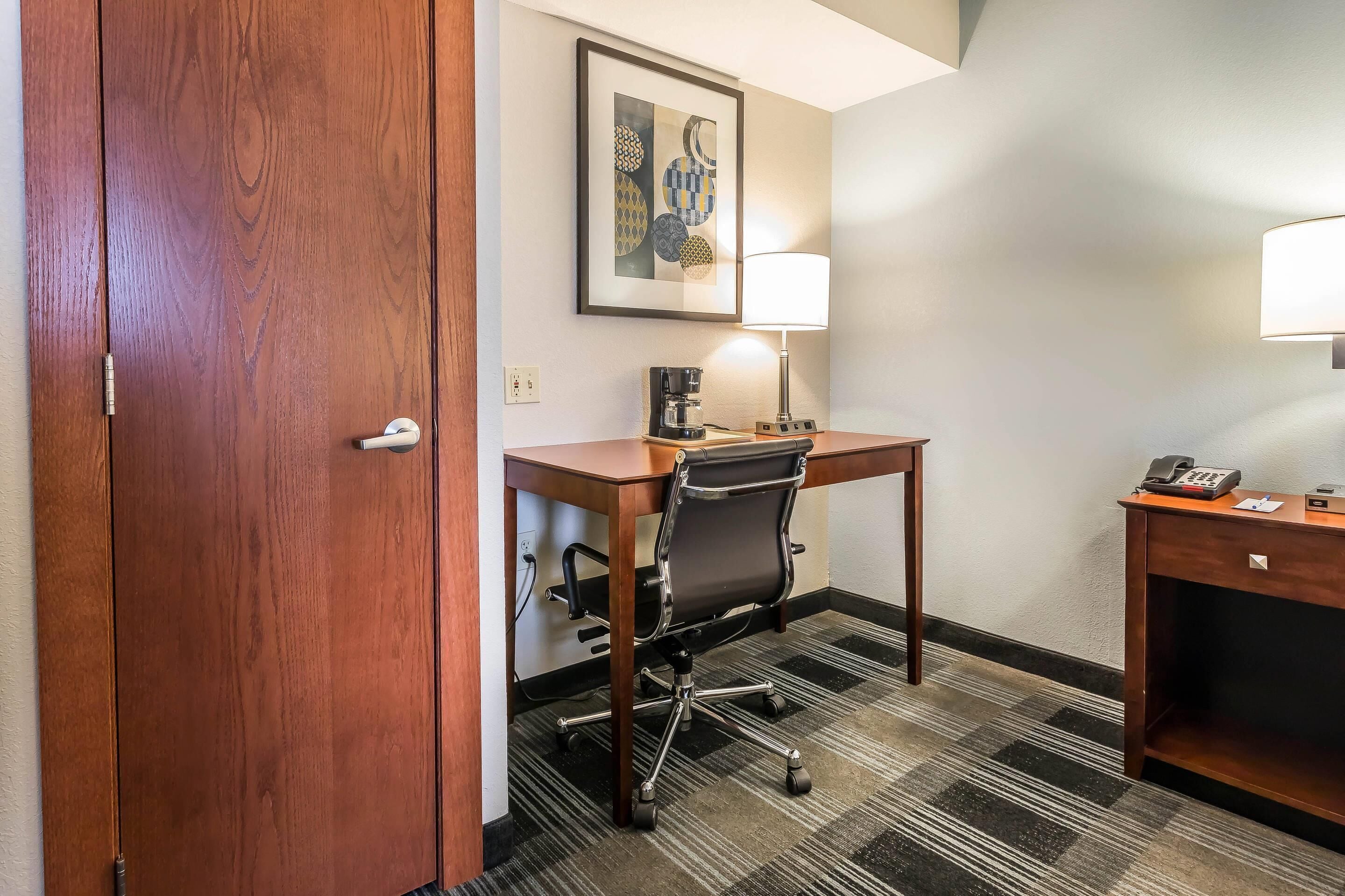 A hotel room with a wooden desk, a chair, a lamp, a coffee maker, and a phone on a small table.