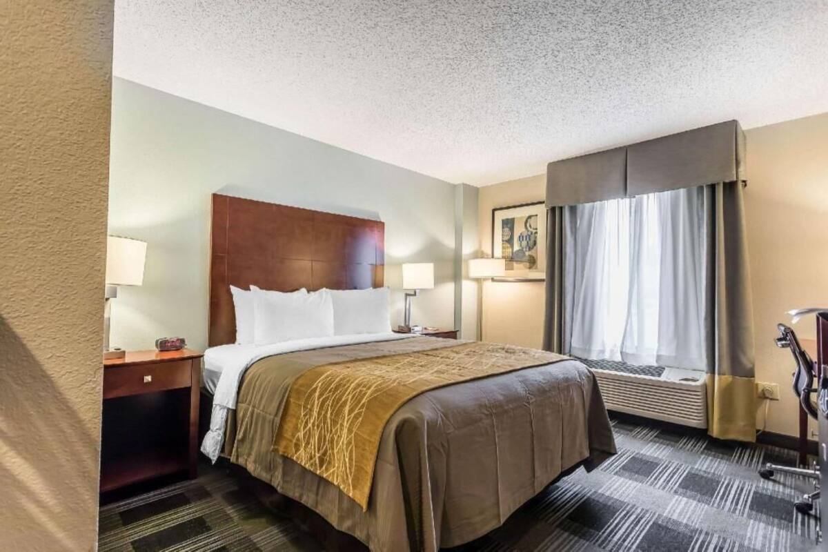 A hotel room with a bed, bedside tables, a desk, a chair, a window with curtains, and a wall-mounted air conditioning unit.