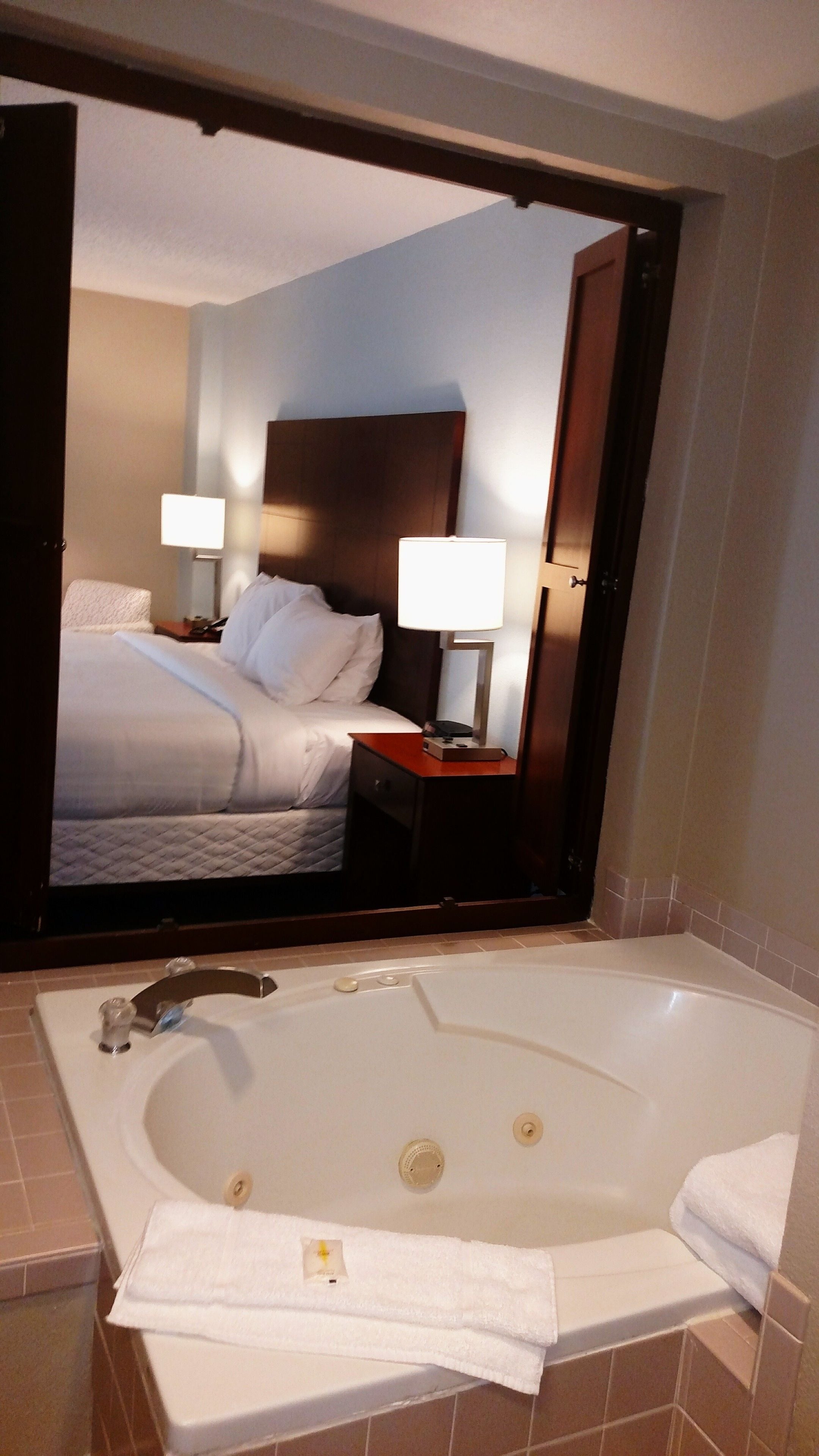 A hotel room with a large bed, two bedside lamps, a nightstand, and a bathtub with a towel.