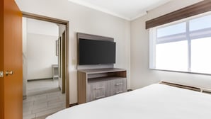 Une chambre à coucher comprenant un lit, un téléviseur fixé sur un meuble en bois, une fenêtre avec des stores et une porte donnant sur une autre pièce.