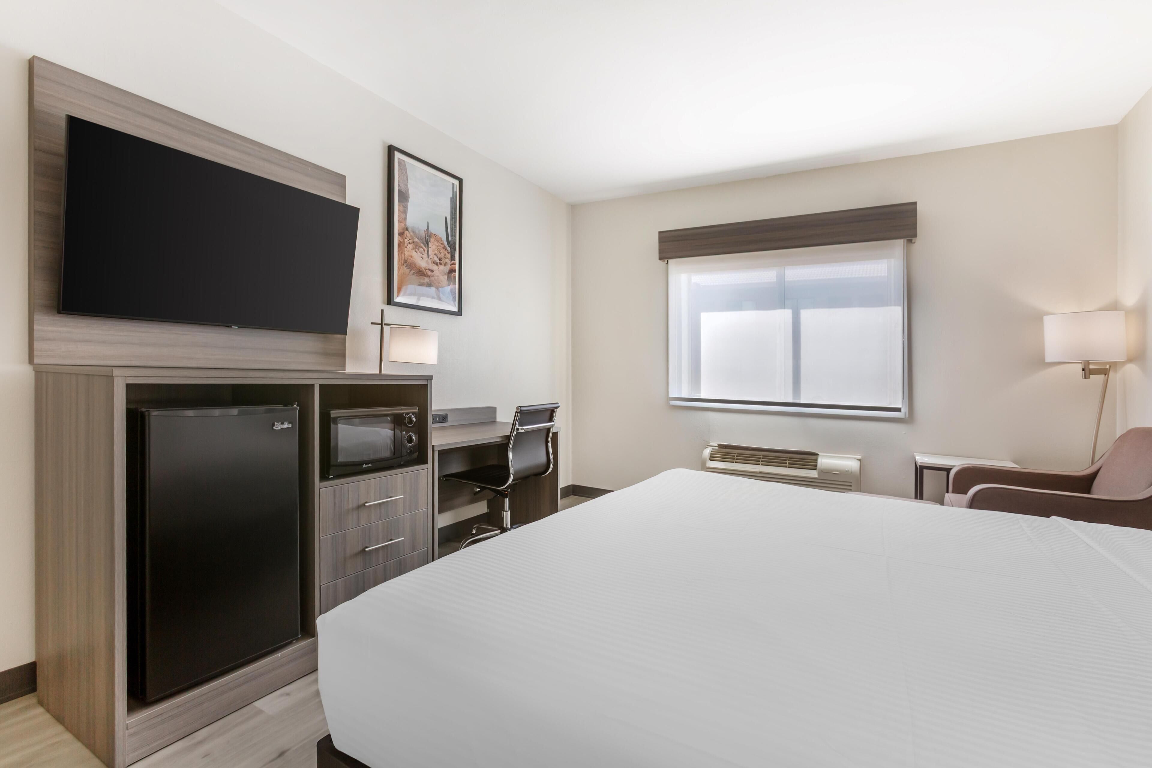 Una camera d'albergo con un letto, una TV a schermo piatto montata su un mobile in legno, una scrivania con una sedia, una finestra con persiane e una lampada fissata al muro.