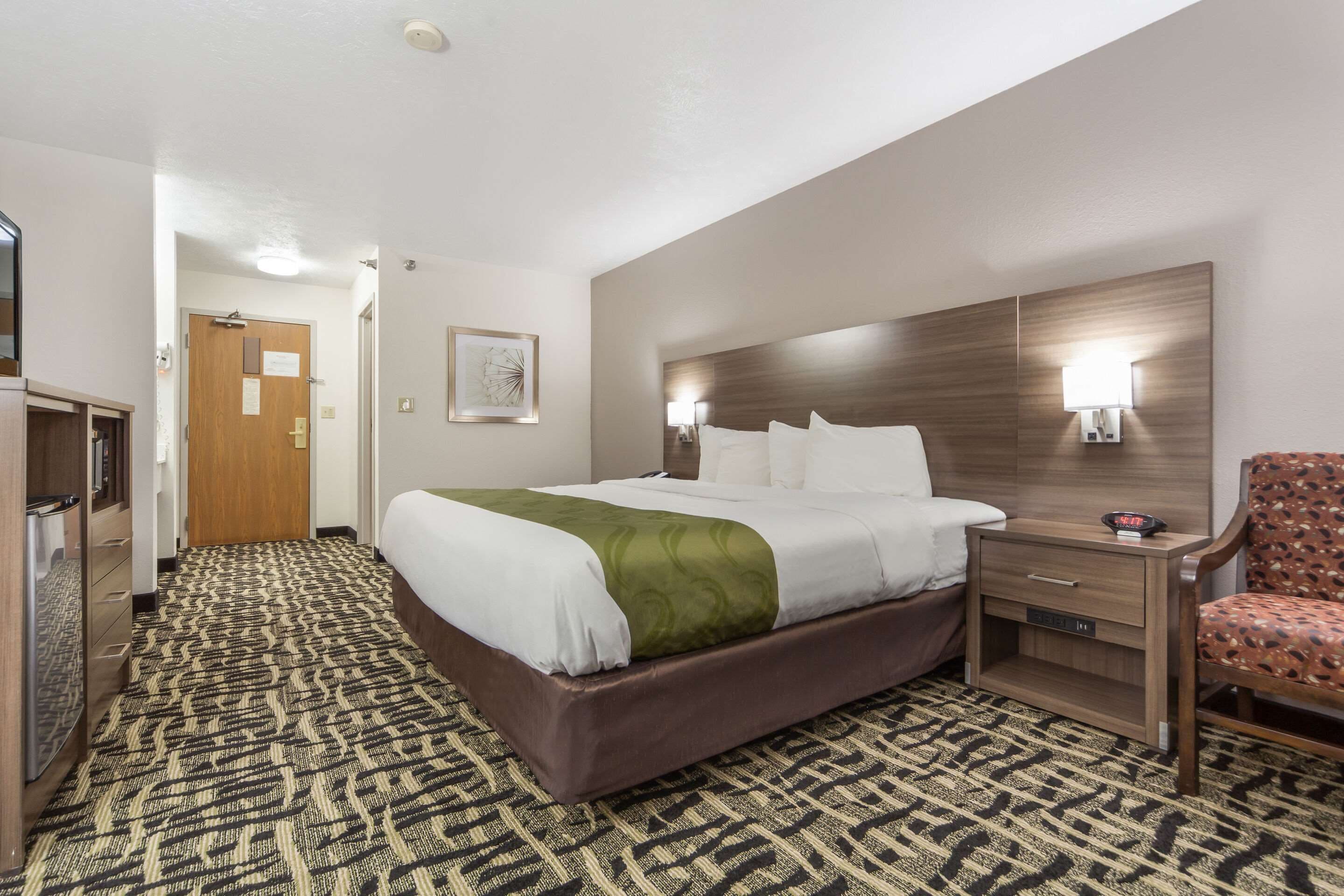 A hotel room with a large bed, a wooden nightstand, a chair, and a patterned carpet.
