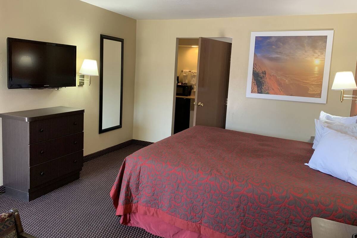 A hotel room with a bed, a television, a dresser, a door leading to a bathroom, and a painting on the wall.