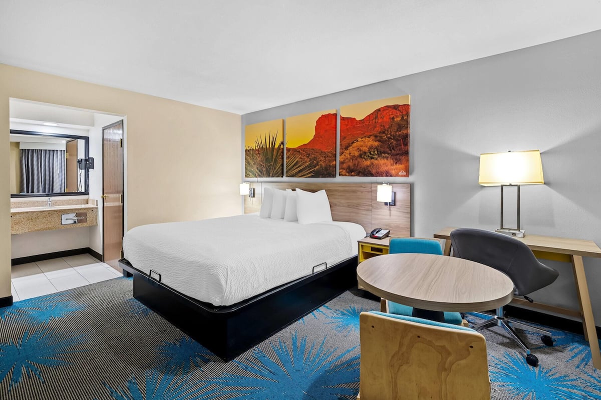 A hotel room with a bed, a desk, a chair, a small table, a mirror, and a painting on the wall.