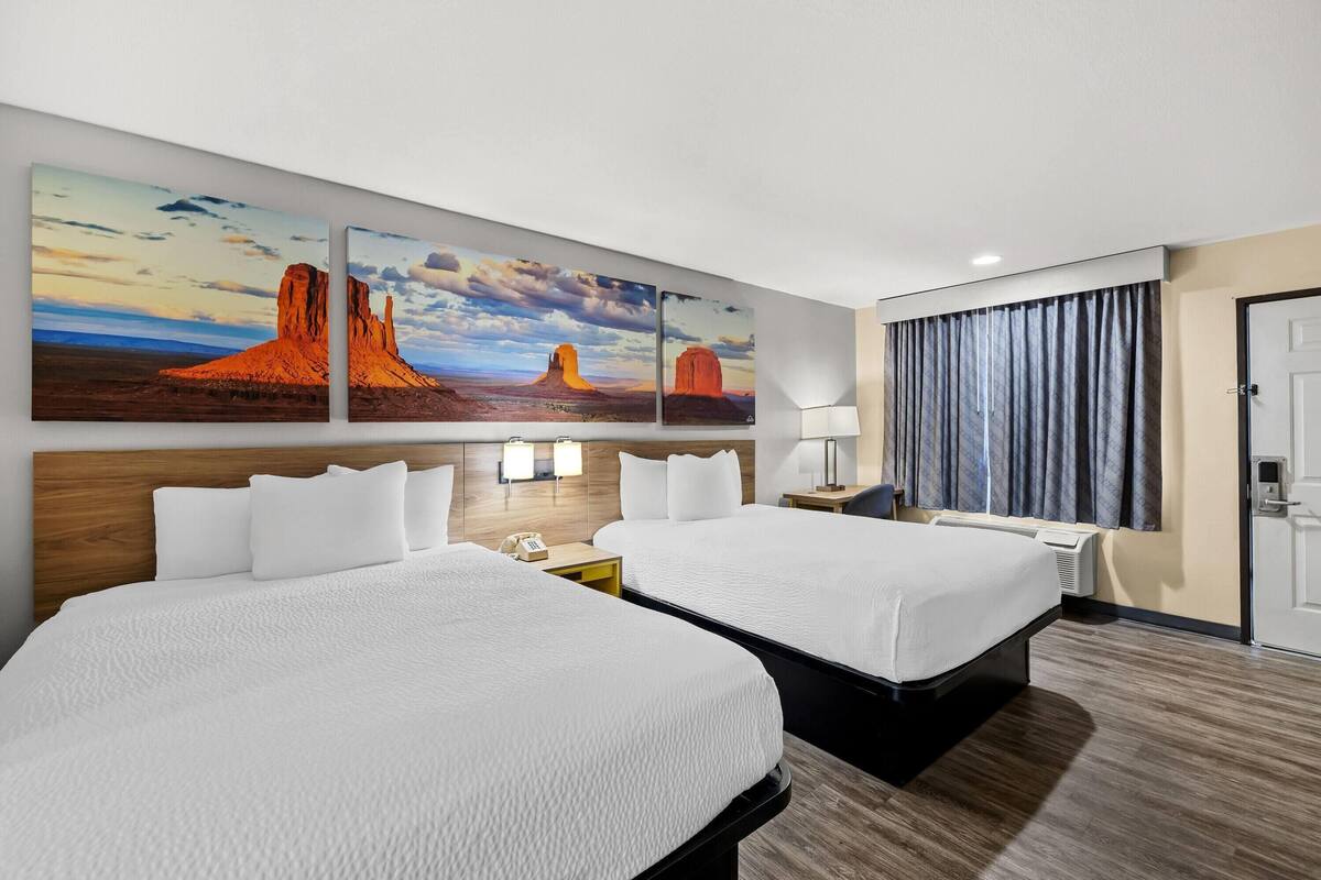 A hotel room with two beds, a large wall mural, a wooden headboard, a bedside table with a lamp, and a door with curtains.
