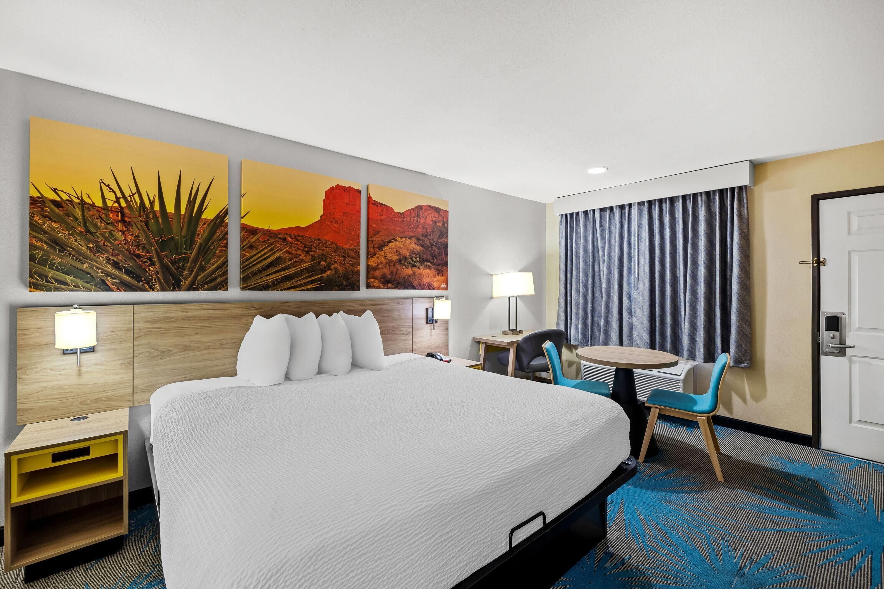 A hotel room with a large bed, a desk with a chair, a small table with a chair, a lamp, and a door.