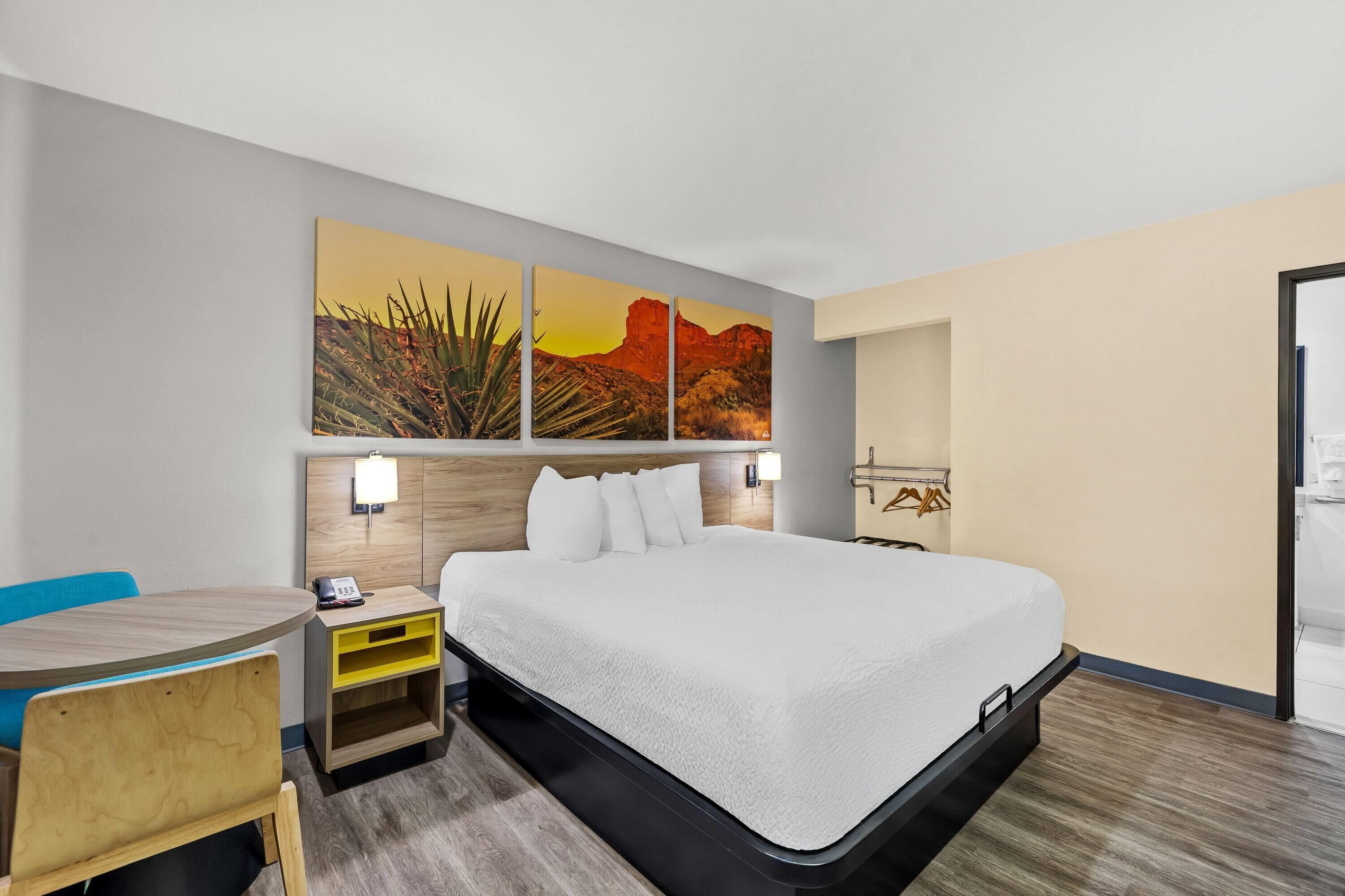 A hotel room with a bed, a desk, and a chair. There are wall-mounted lights and a painting above the bed.