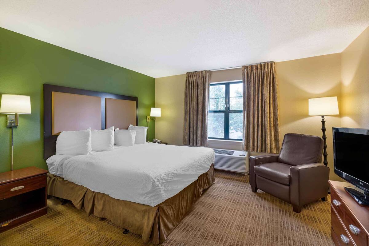 A hotel room with a large bed, two bedside tables, a flat-screen TV, a chair, and a window with curtains.