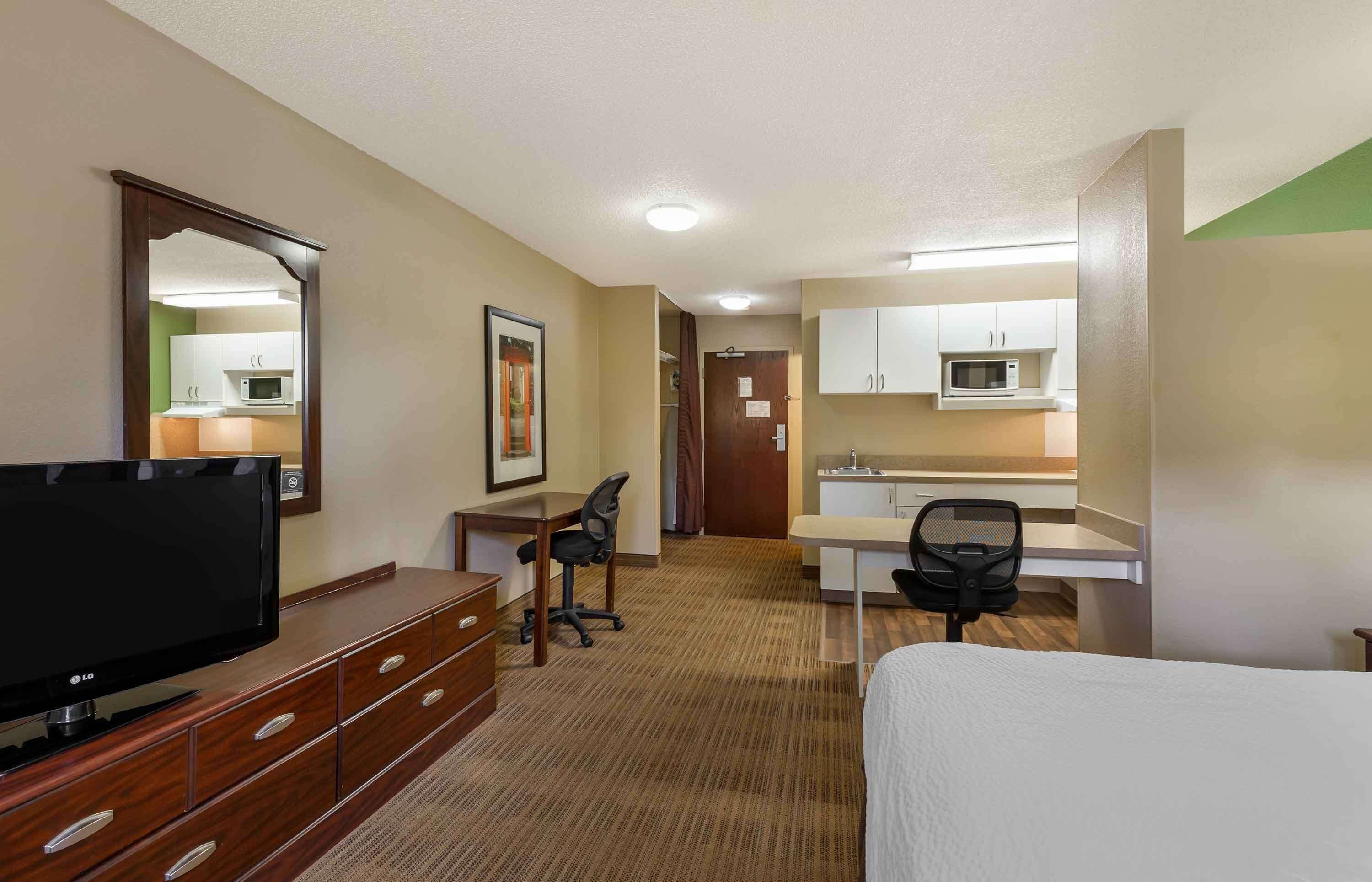 A hotel room with a bed, a desk, a chair, a TV, a dresser, and a kitchenette.