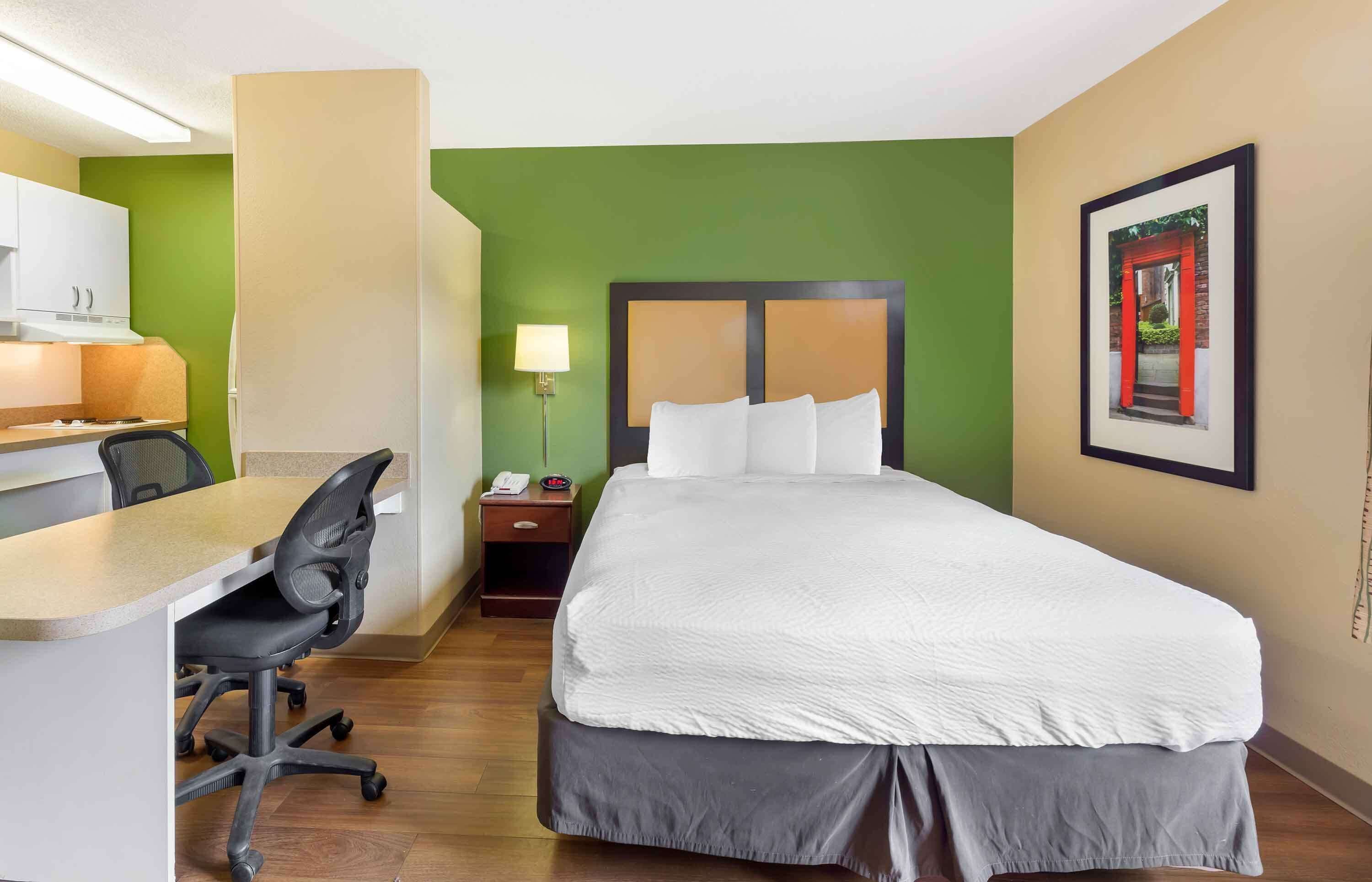 A hotel room with a bed, desk, chair, and a nightstand with a lamp.