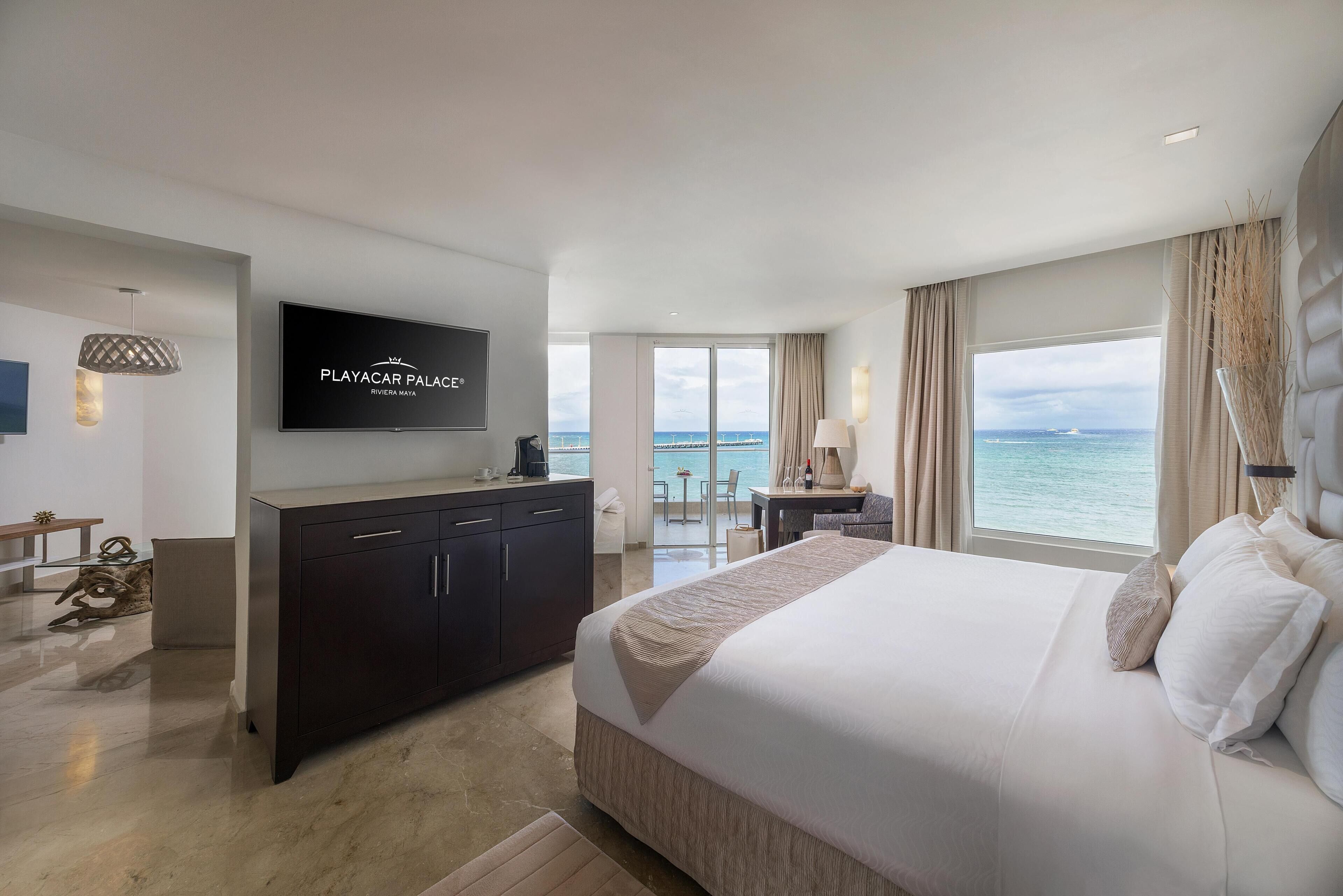 Quarto de hotel moderno com uma cama grande, televisão fixada na parede, escrivaninha com cadeira e vista para o oceano.
