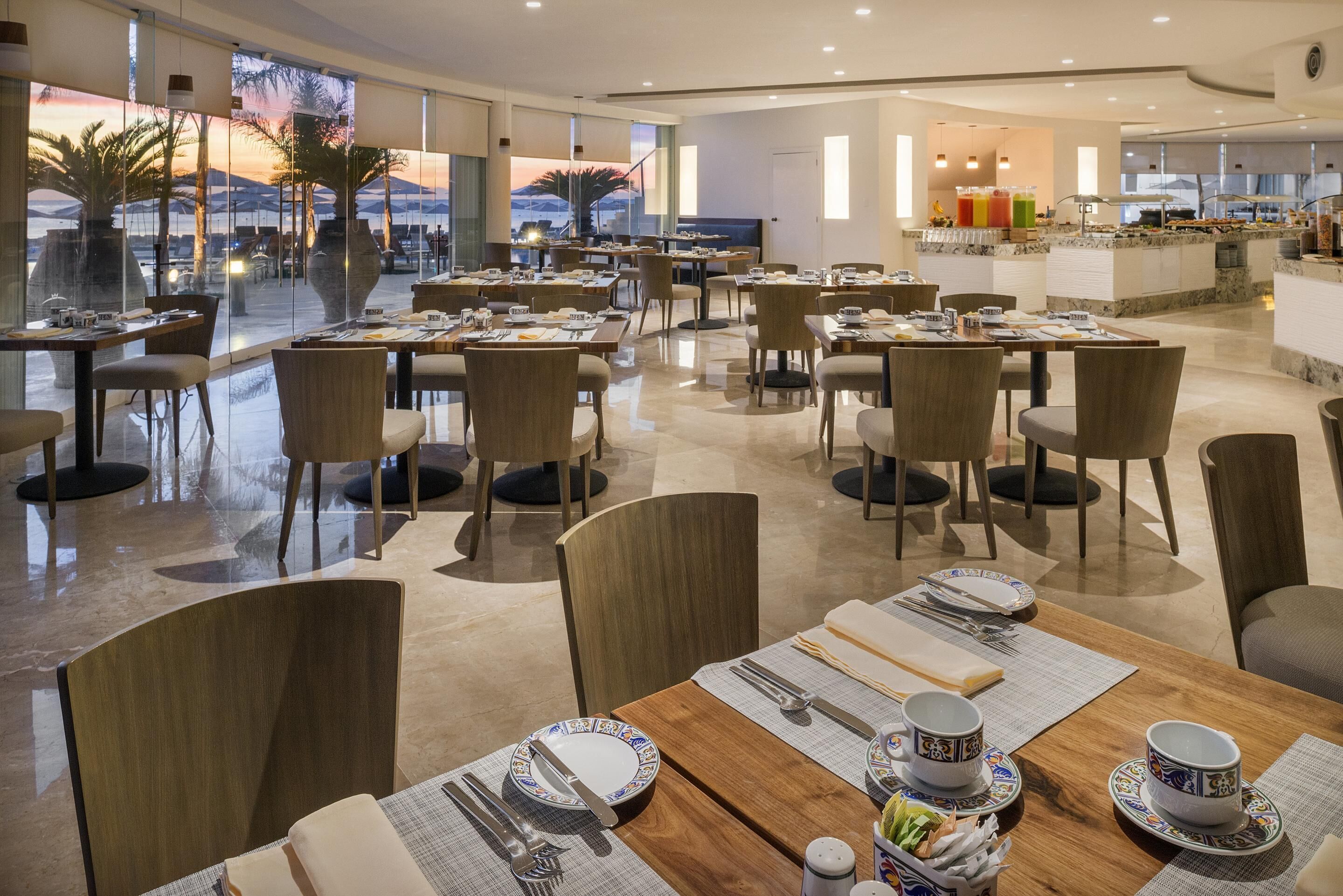 A spacious dining area with multiple tables and chairs, large windows offering a view of palm trees and mountains, and a buffet area in the background.
