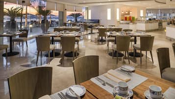 A spacious dining area with multiple tables and chairs, large windows offering a view of palm trees and mountains, and a buffet area in the background.