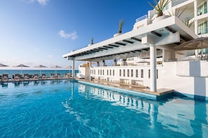 A pool area with lounge chairs and umbrellas, a white building with balconies, and a clear view of the sea. - Playacar Palace All Inclusive (Playa del Carmen)