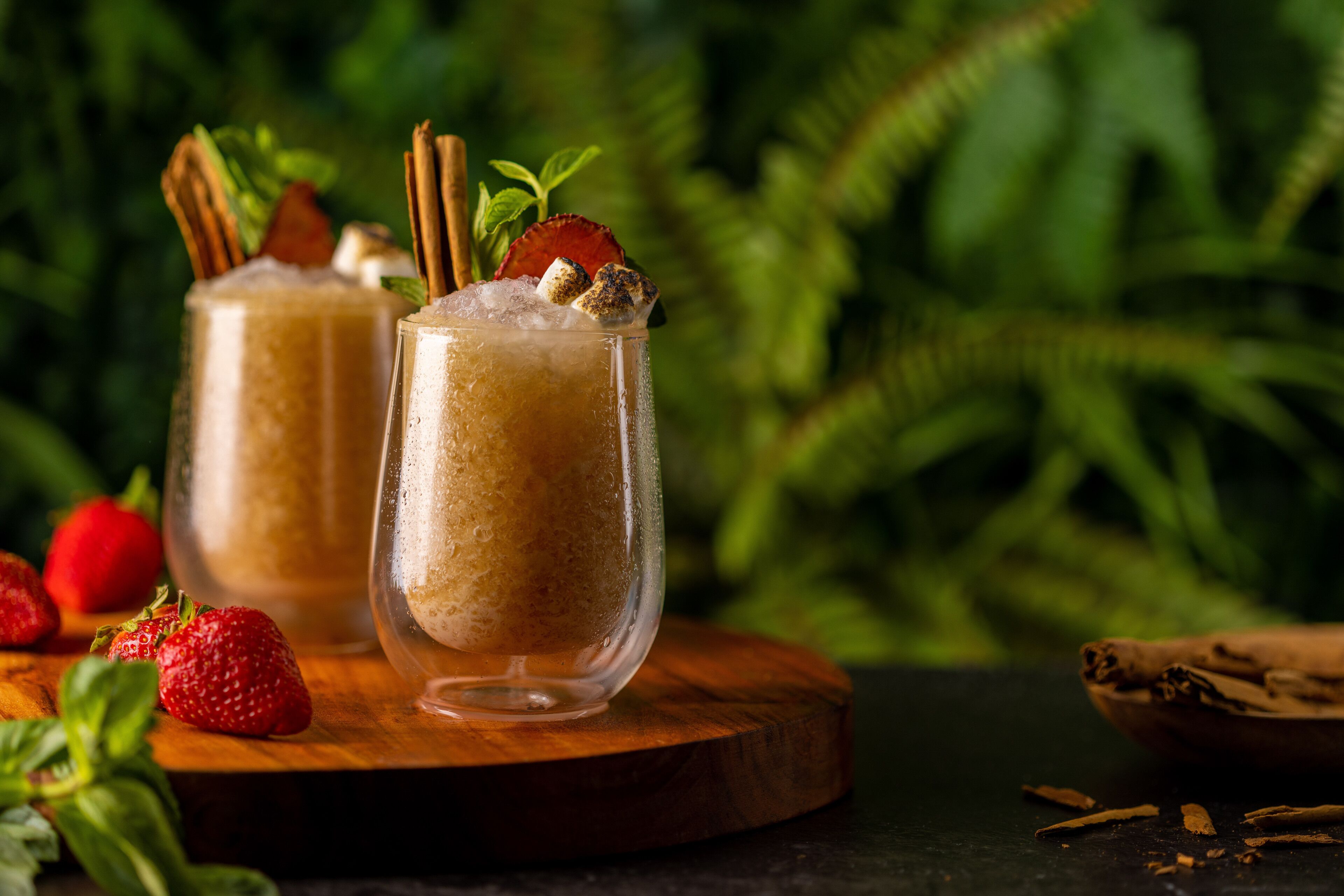 Two glasses of a frothy beverage with strawberries and cinnamon sticks, placed on a wooden board with a dark background and green foliage.
