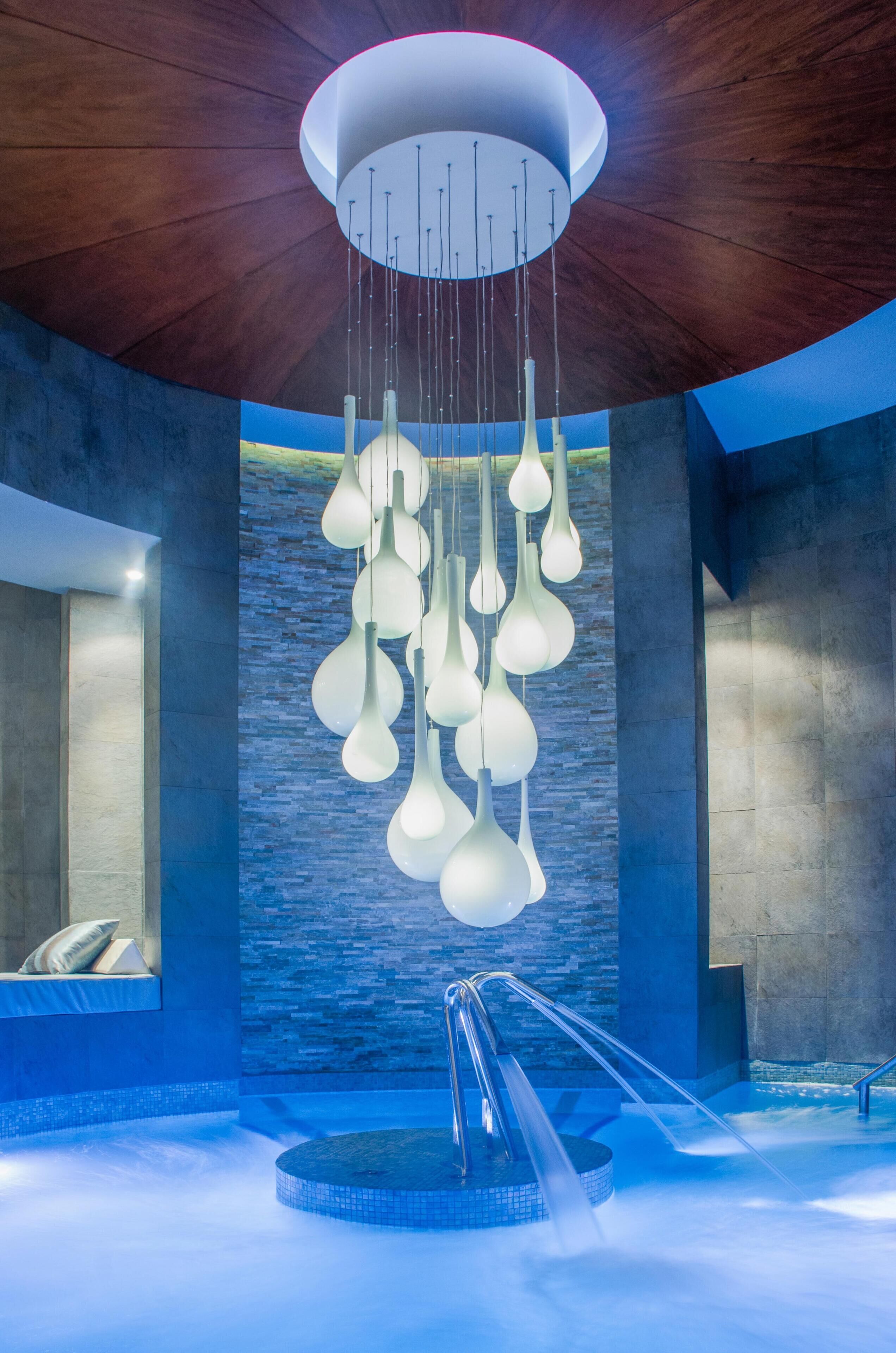 Un espace piscine intérieur moderne, doté d’un plafond au design original, d’un élément décoratif aquatique et d’un mur en pierre en arrière-plan.