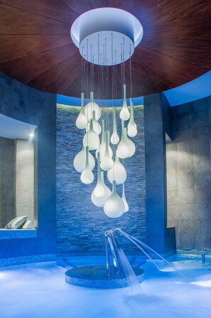 A modern indoor pool area with a unique ceiling design, a water feature, and a stone wall backdrop.
