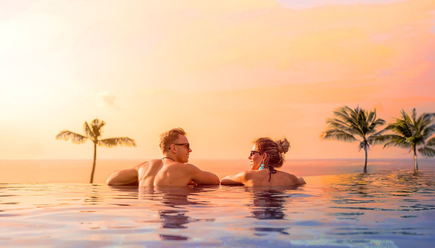 A couple in a private pool with palm trees and a sunset in the background.
