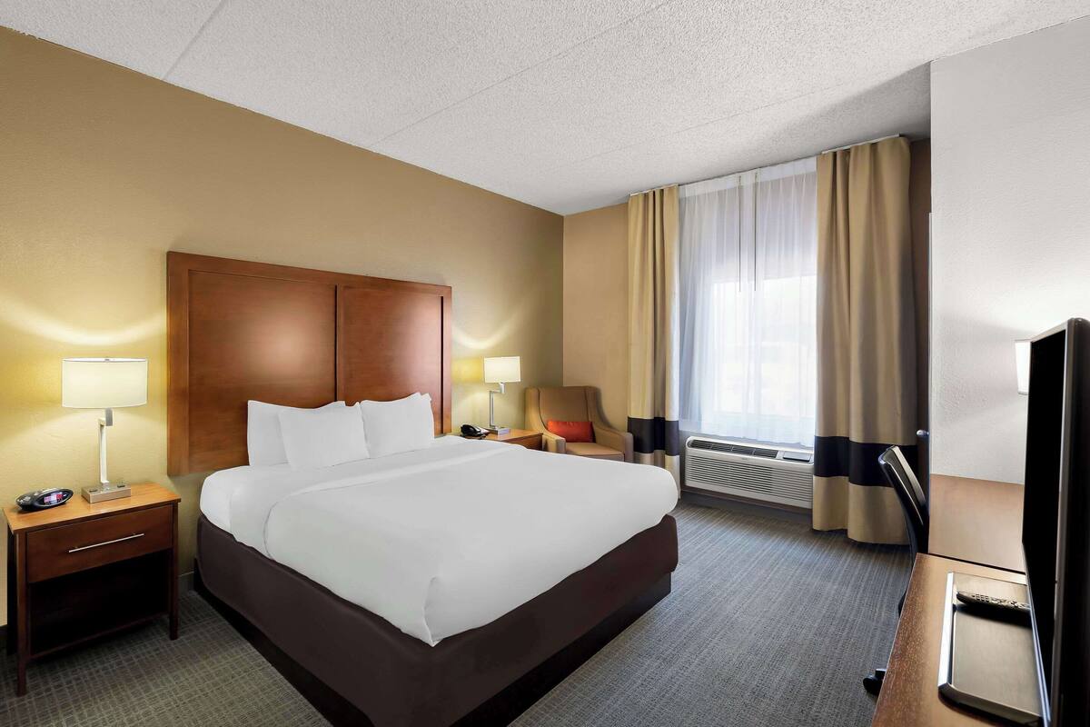 A hotel room with a large bed, two bedside tables with lamps, a chair, a desk with a TV, and a window with curtains.