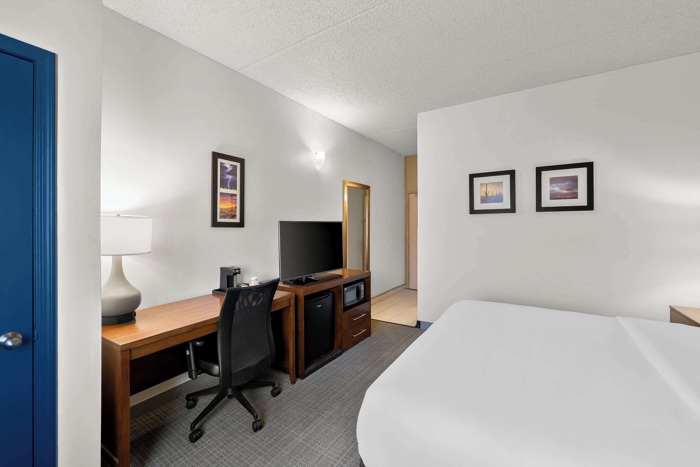 A hotel room with a bed, a desk with a computer, a television, a mirror, and two framed pictures on the wall.