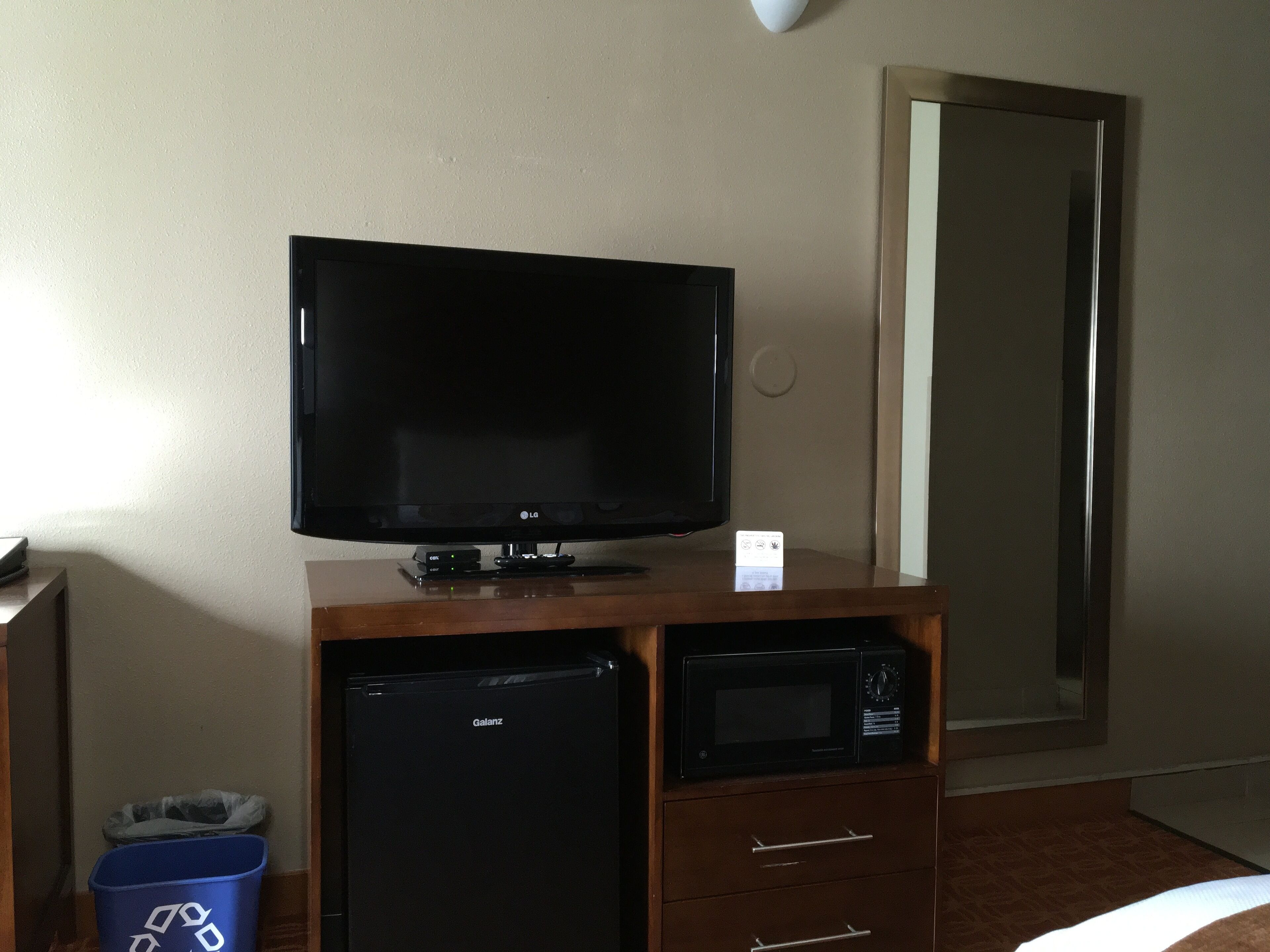 Une chambre d’hôtel équipée d’un téléviseur à écran plat posé sur un meuble en bois, d’un petit réfrigérateur noir, d’un four à micro-ondes et d’une poubelle de recyclage bleue.