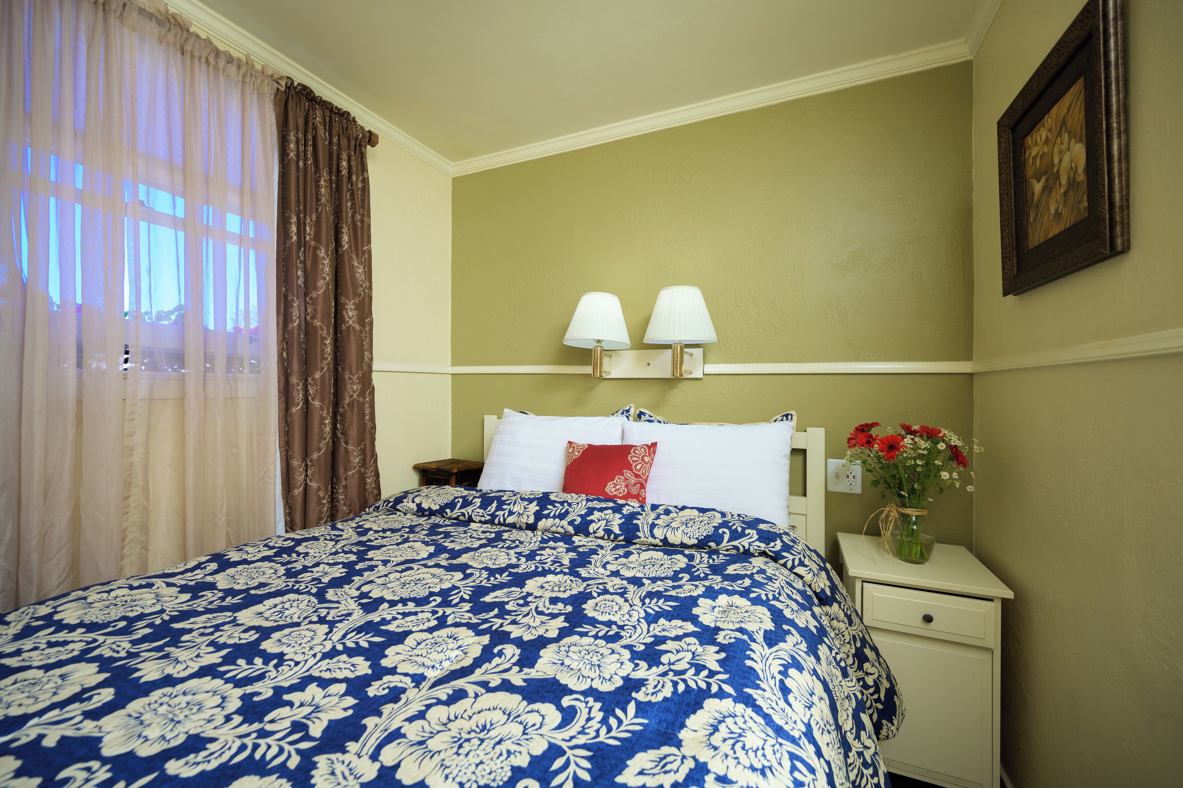 Una cama bien tendida con una colcha azul y blanca de motivos florales, una mesita de noche con un jarrón de flores y una lámpara fijada a la pared.