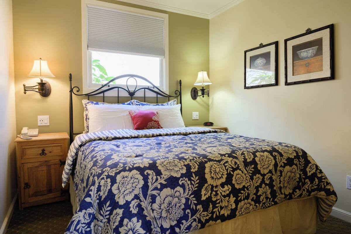 A bedroom with a bed, nightstands, wall-mounted lamps, a window with blinds, and two framed pictures on the wall.