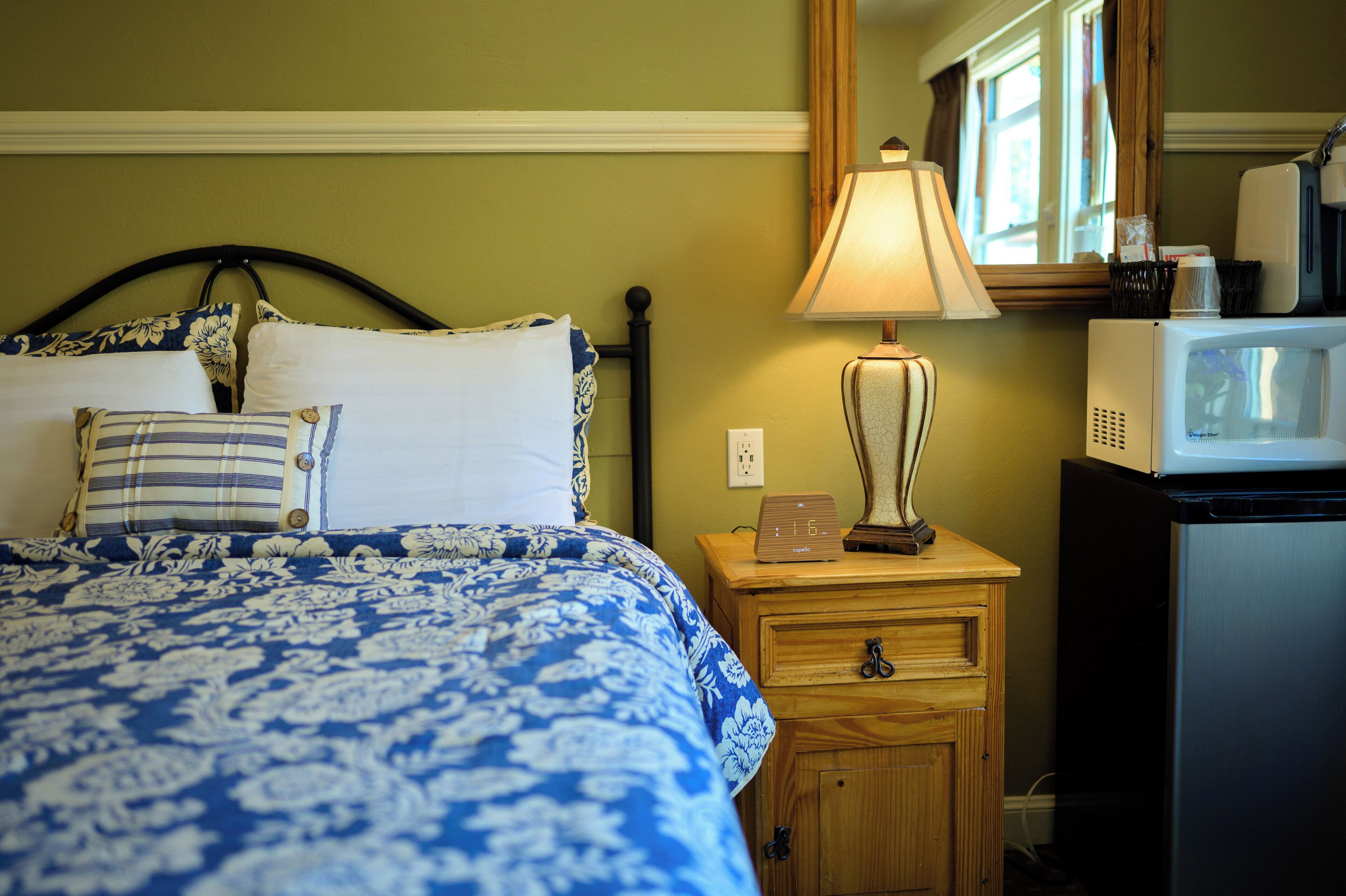 A neatly made bed with a blue and white floral bedspread, a wooden nightstand with a lamp, and a microwave on a black cabinet.