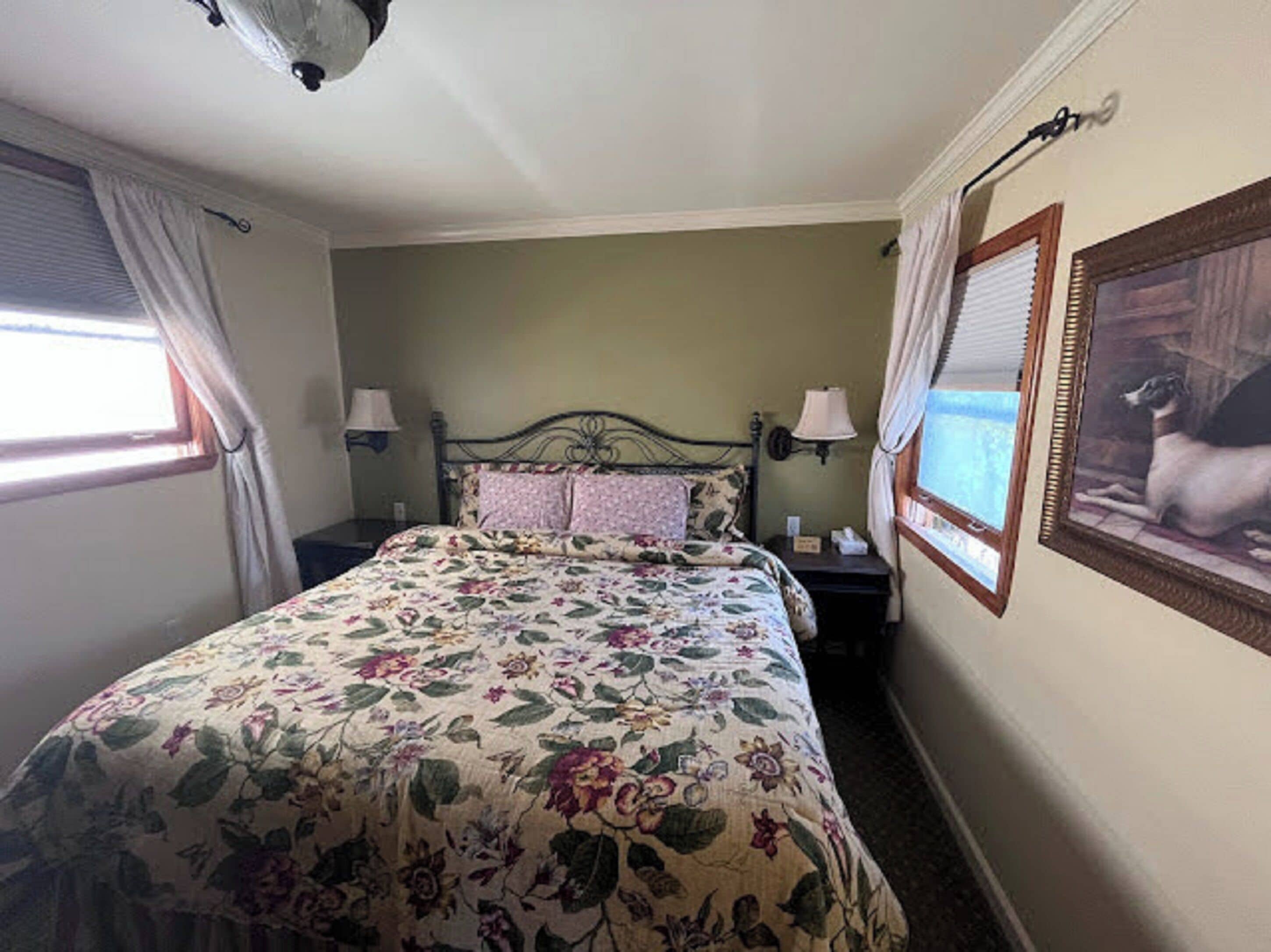 Een slaapkamer met een bloemenbedovertrek, een nachtkastje met een lamp, een wandlamp en een lijst met een foto van een hond.