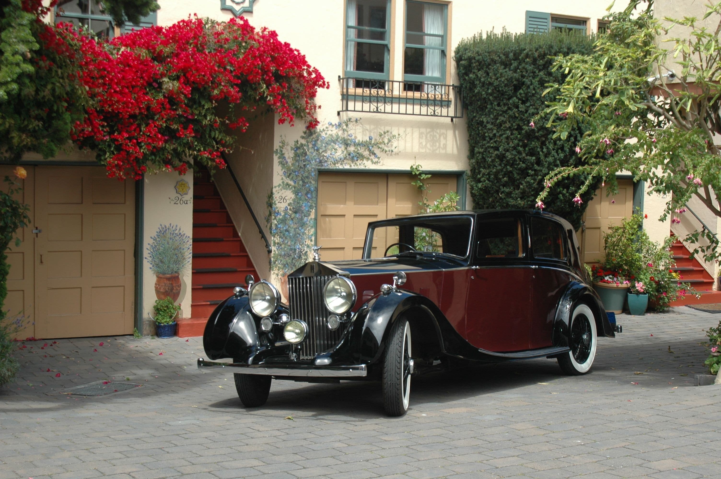 Un coche antiguo estacionado frente a un edificio con puerta de garaje, escalones y plantas en macetas.