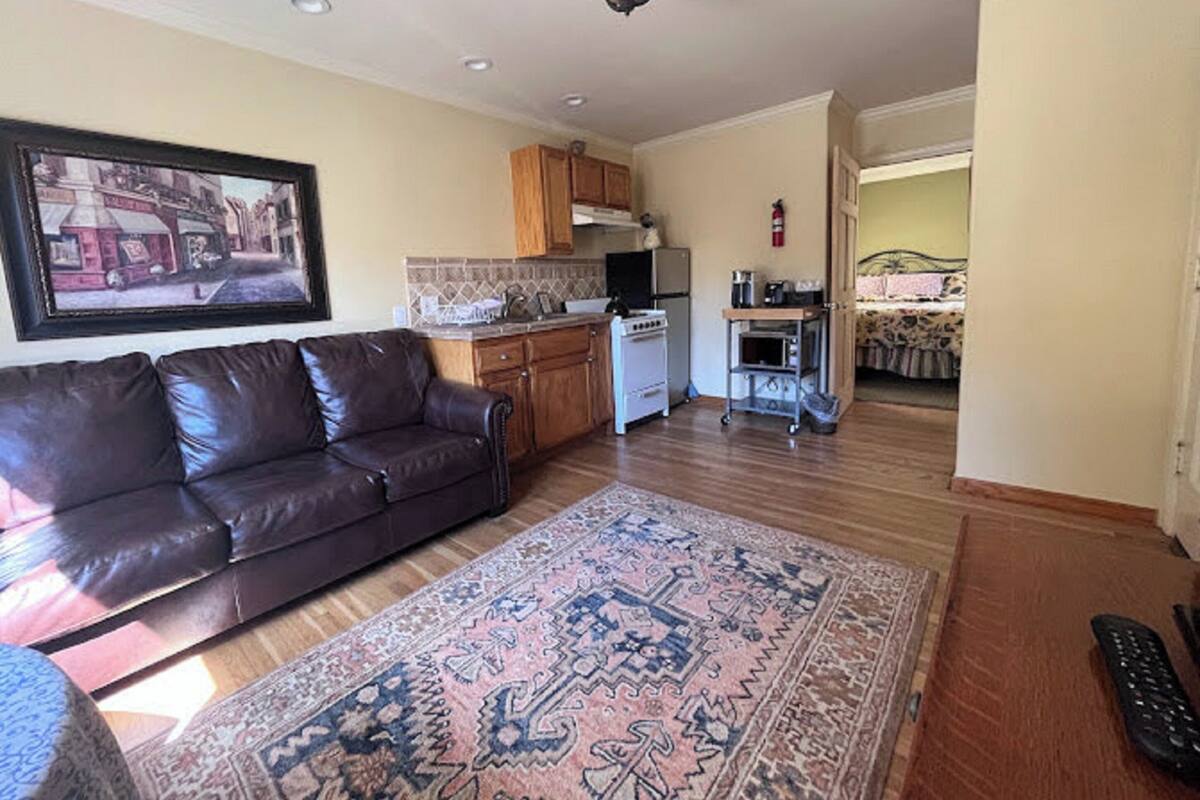 A living room with a brown leather sofa, a patterned rug, a kitchen with wooden cabinets, and a dining area with a table and chairs.