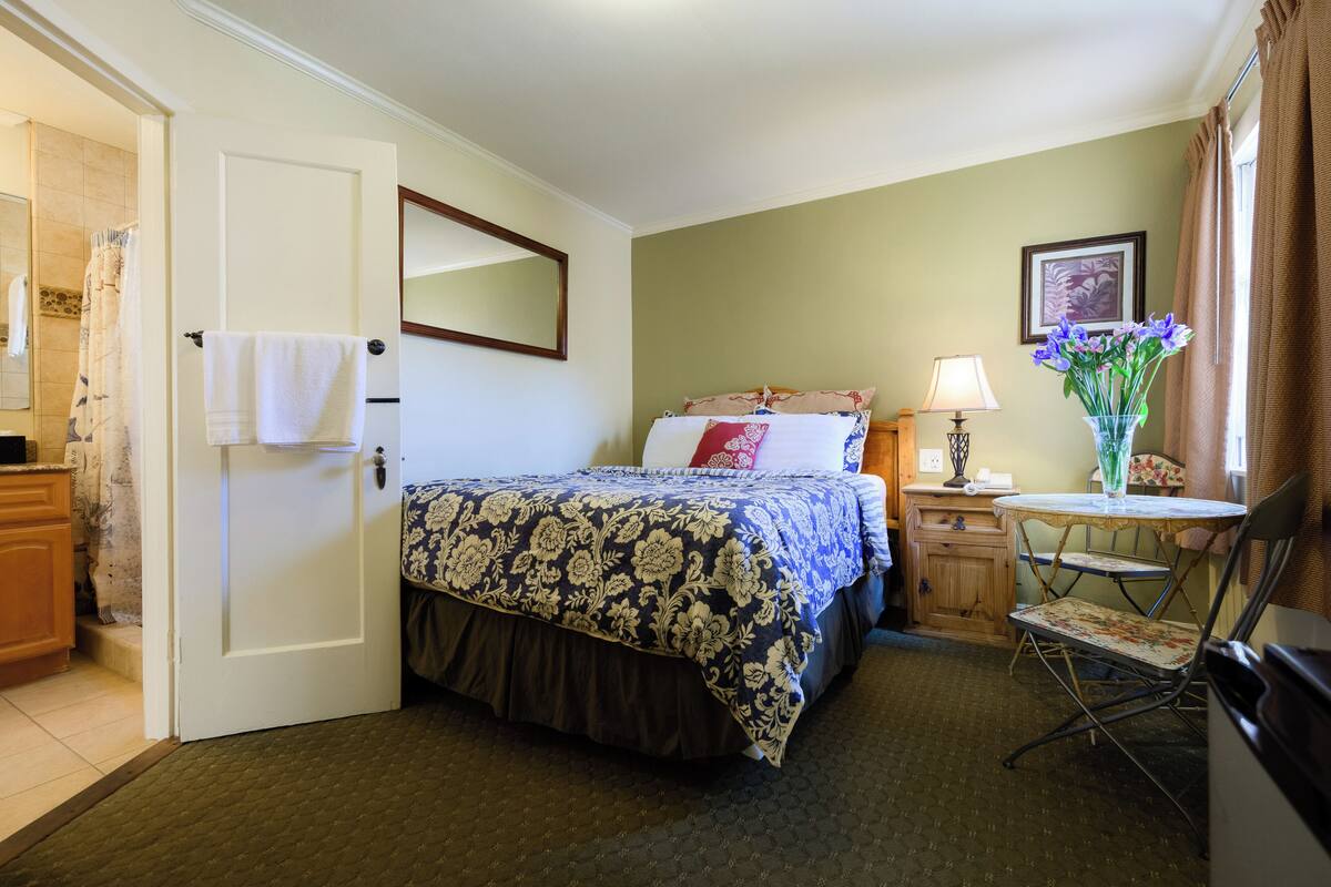 A hotel room with a bed, a chair, a table, a lamp, and a vase with flowers.