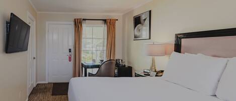 Ein Hotelzimmer mit einem Bett, einem Schreibtisch mit Stuhl, einem an der Wand befestigten Fernseher, einem Fenster mit Vorhängen und einem gerahmten Bild an der Wand.