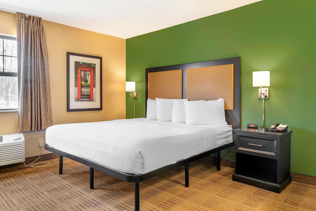 Une chambre d’hôtel avec un grand lit, deux tables de chevet, une commode, un tableau encadré au mur et une fenêtre avec des rideaux.