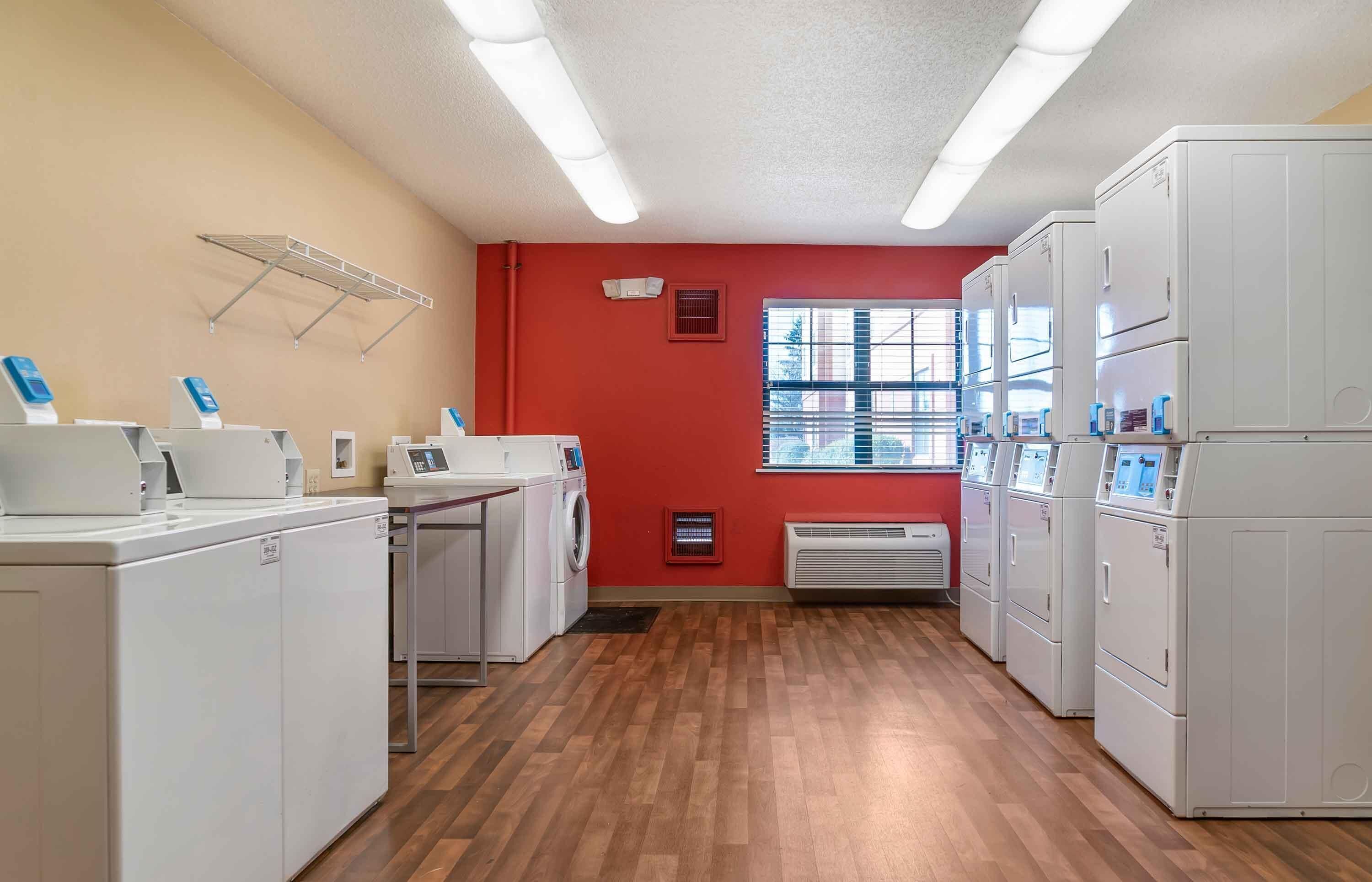A laundry room with white appliances, a red wall, and a window with blinds.