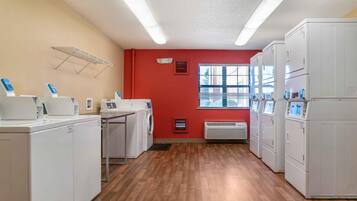 A laundry room with white appliances, a red wall, and a window with blinds.