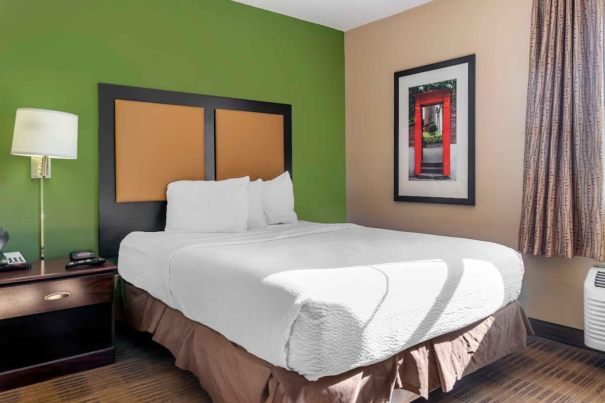 Une chambre d’hôtel avec un lit, une table de chevet avec une lampe, un tableau encadré au mur et une fenêtre avec des rideaux.