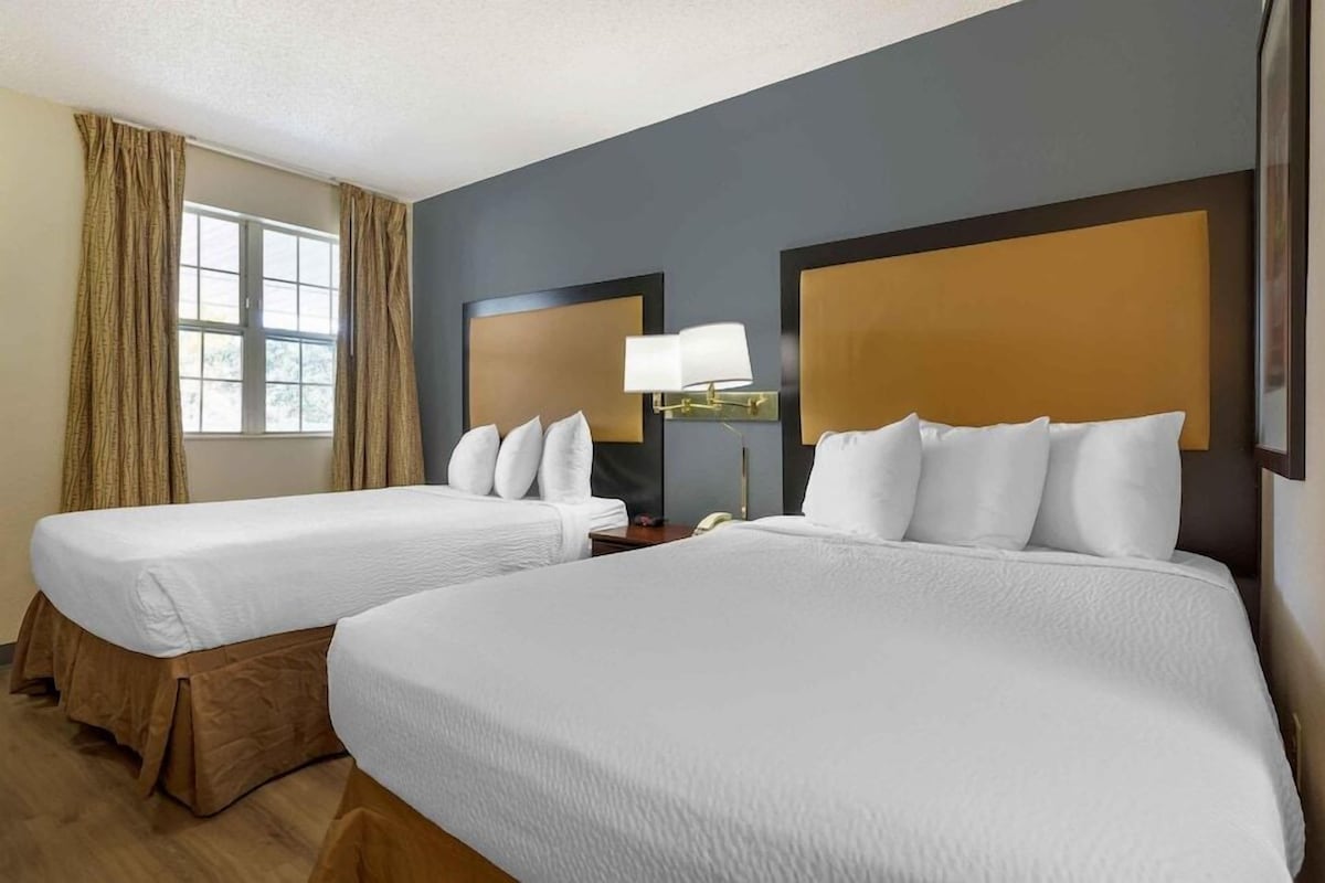 A hotel room with two beds, a window with curtains, a bedside lamp, and a wall-mounted light fixture.