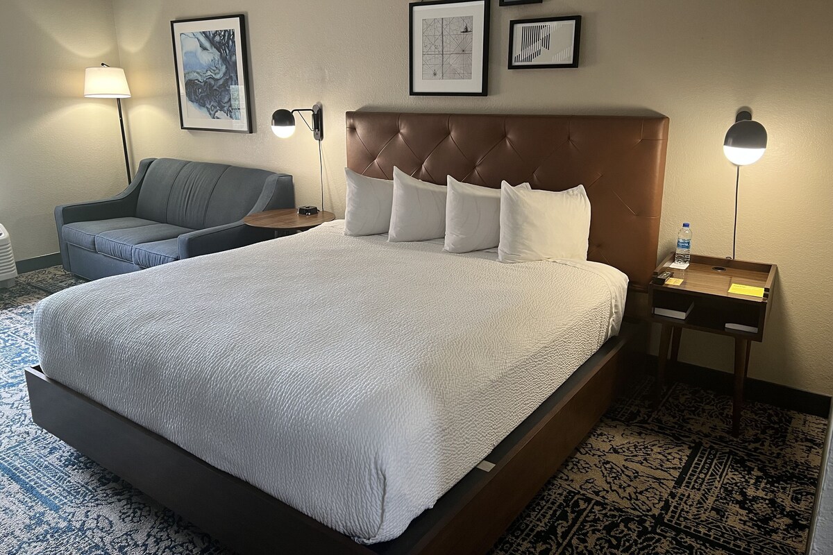 A hotel room with a large bed, a sofa, a nightstand with a lamp, and framed artwork on the wall.