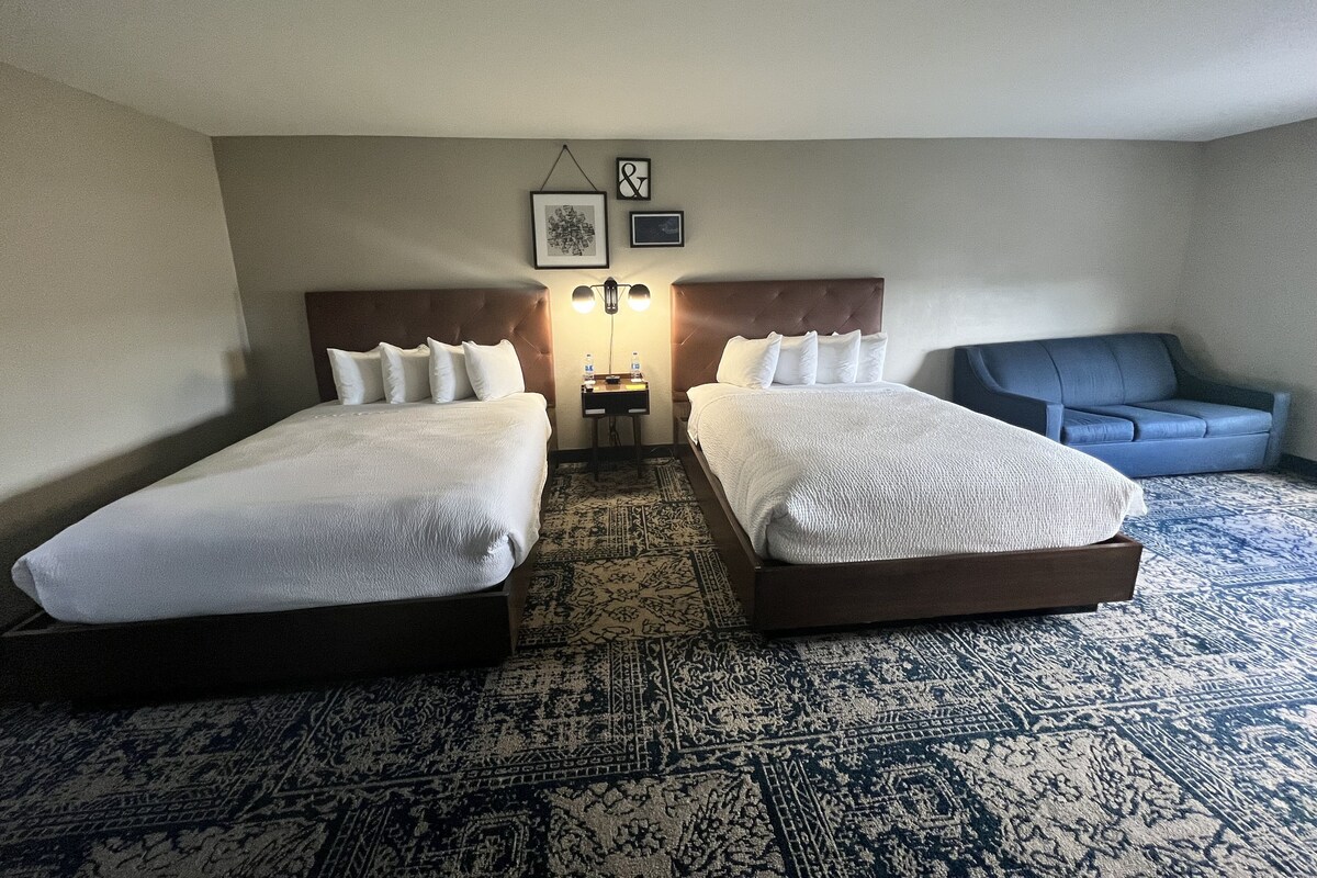 A hotel room with two beds, a nightstand, a lamp, a blue sofa, and a patterned rug.