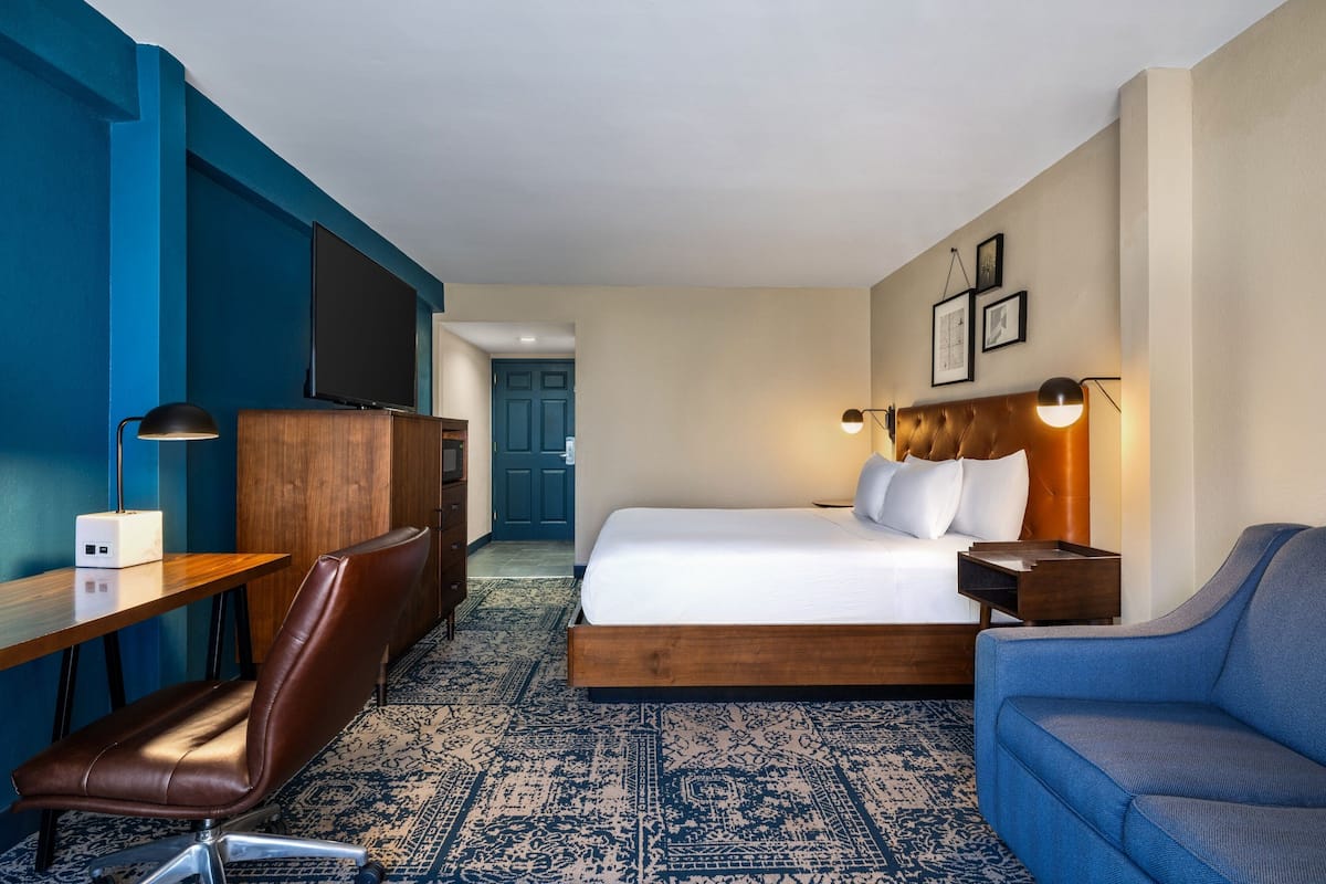 A hotel room with a bed, a desk, a chair, a sofa, a TV, and a lamp.