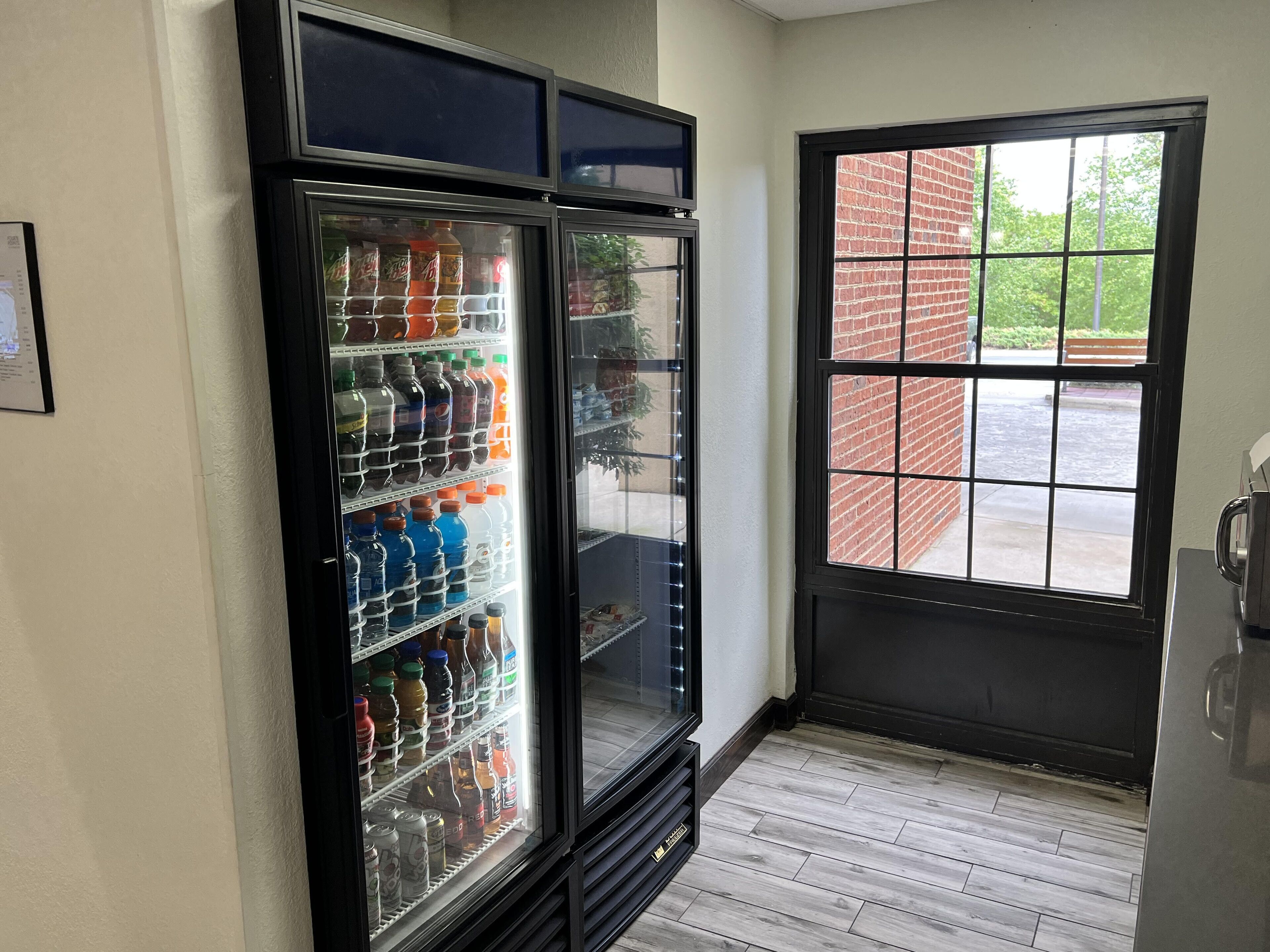 A vending machine stocked with beverages and a window with a view of a brick building and greenery.