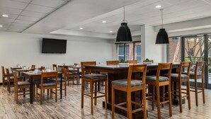 A modern dining area with wooden tables and chairs, a large flat-screen TV mounted on the wall, and pendant lighting.