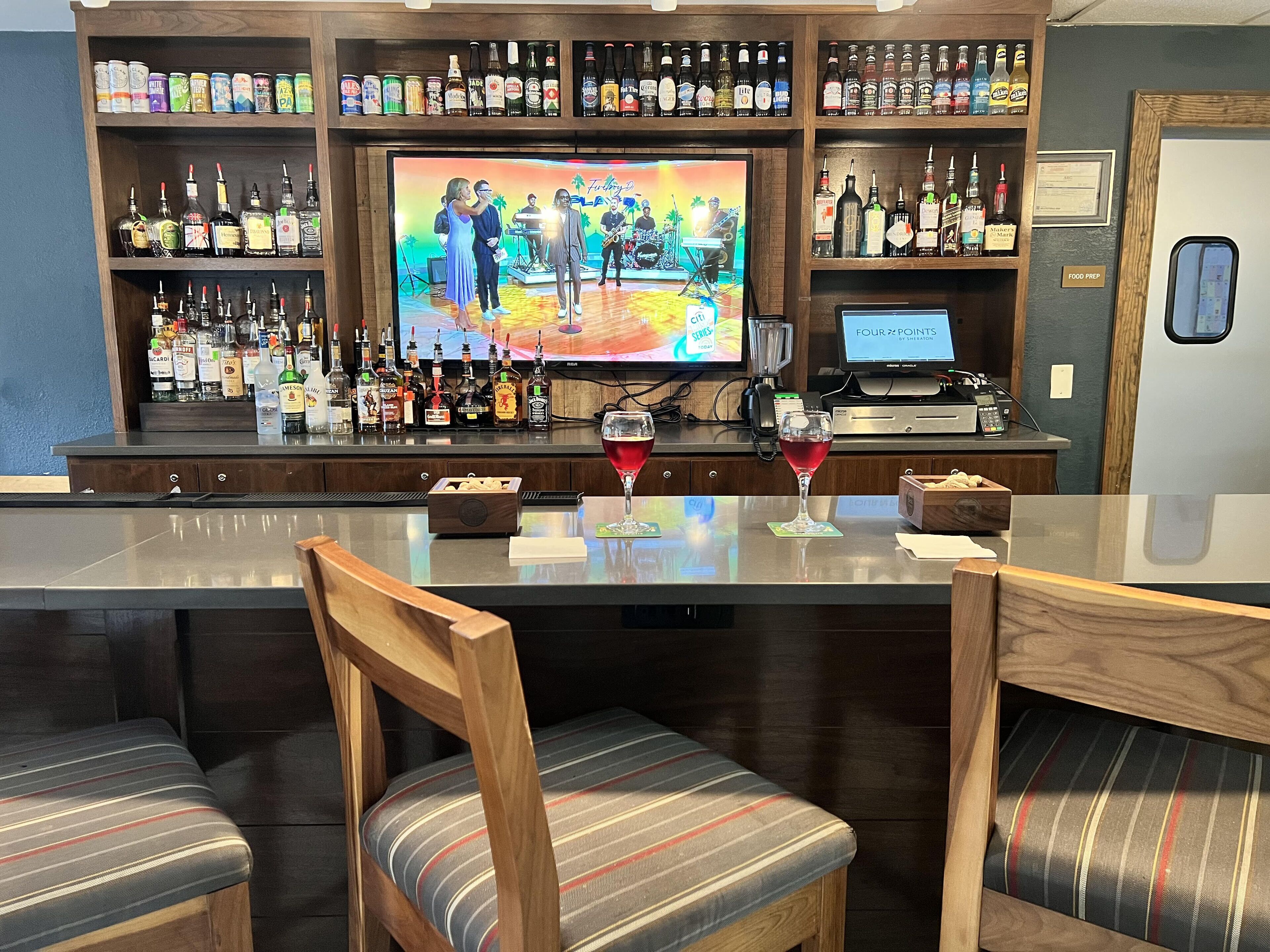 Zona de bar con una televisión mostrando una presentación de banda, estantes con botellas de licor y una barra con dos copas de vino tinto.